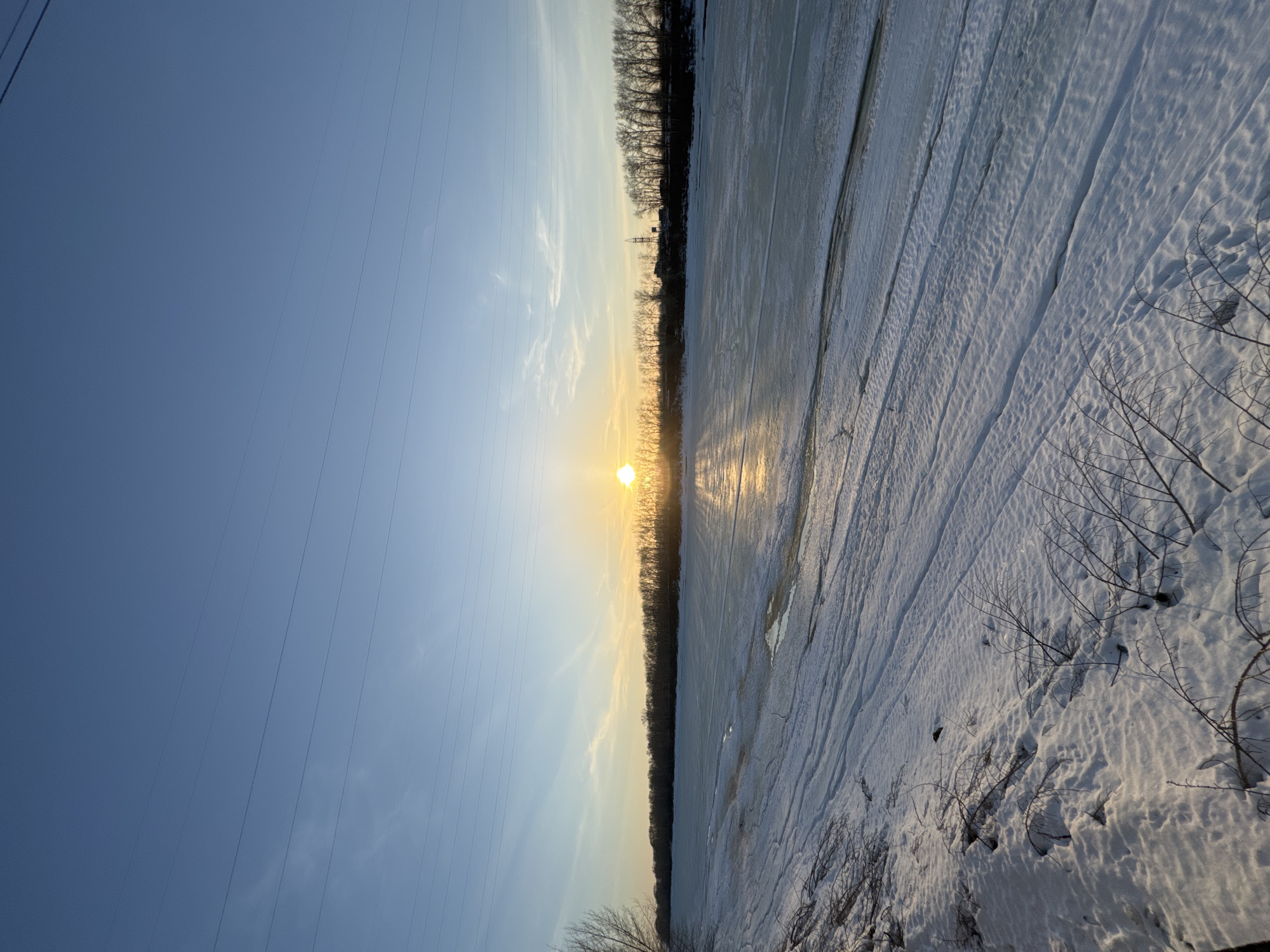The photograph shows a winter landscape at sunset. The sun, low on the horizon, is positioned between a treeline and reflects light onto an ice-covered surface, likely a frozen river or lake. Tire tracks are visible in the ice. To the left, utility lines stretch across a light blue sky. Sparse vegetation, possibly bushes or small trees, are visible in the lower right corner. The specific location within Chaikovsky, Russia, cannot be determined solely from the visual content of the image.