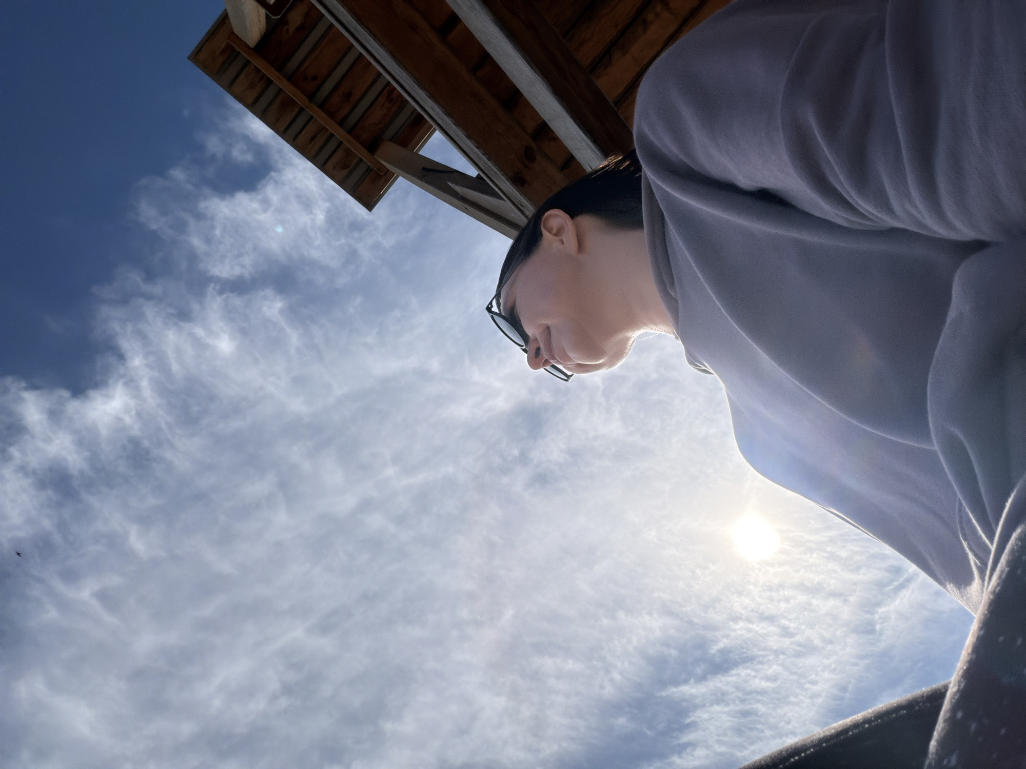 This image is an upward-angled shot capturing a person, a wooden structure, and a bright, cloudy sky. The perspective is from below, looking up.
A person is prominently featured on the right side of the frame, seen in profile from the neck up, facing towards the left and slightly downwards. They have short, dark hair and are wearing dark-rimmed glasses. The person is dressed in a light grey, long-sleeved top or sweatshirt, which occupies a significant portion of the right foreground. Their expression appears neutral, and they seem to be looking at something below the camera's view.
The sky dominates the background, displaying a bright blue expanse with numerous scattered, wispy white clouds, characteristic of cirrus or altocumulus formations. The sun is clearly visible in the lower right quadrant, radiating intense light and causing a lens flare effect, indicating a sunny day. A small, dark, bird-like object is visible against the blue sky in the upper left corner.
In the upper left corner of the image, a dark wooden structure is visible. It appears to be the underside of an overhang, possibly a roof, deck, or gazebo, constructed from horizontal wooden planks and supporting beams. This structure provides a stark, dark contrast against the brightly lit sky.
The scene depicts an outdoor setting, likely on a sunny day in Chaikovsky, Russia. The time of day is suggested to be late morning or afternoon due to the sun's bright, slightly lower position. No text is visible in the image.