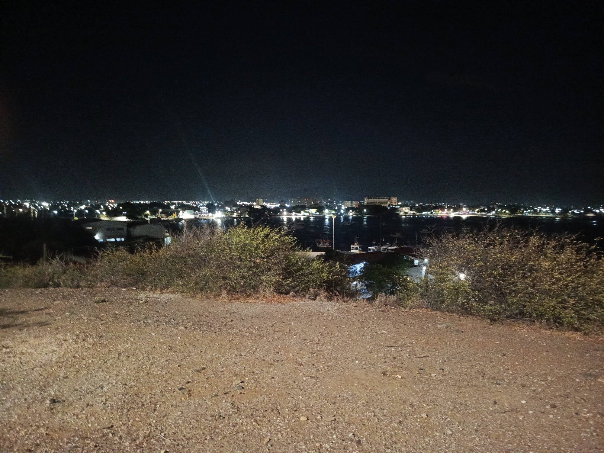 The image shows a night view of Juan Griego, Venezuela. The foreground features a dirt ground and shrubs. In the distance, the city lights are visible along the coastline, reflected in the water. The scene appears to be taken from an elevated position overlooking the city and the bay. There are no people visible in the image.