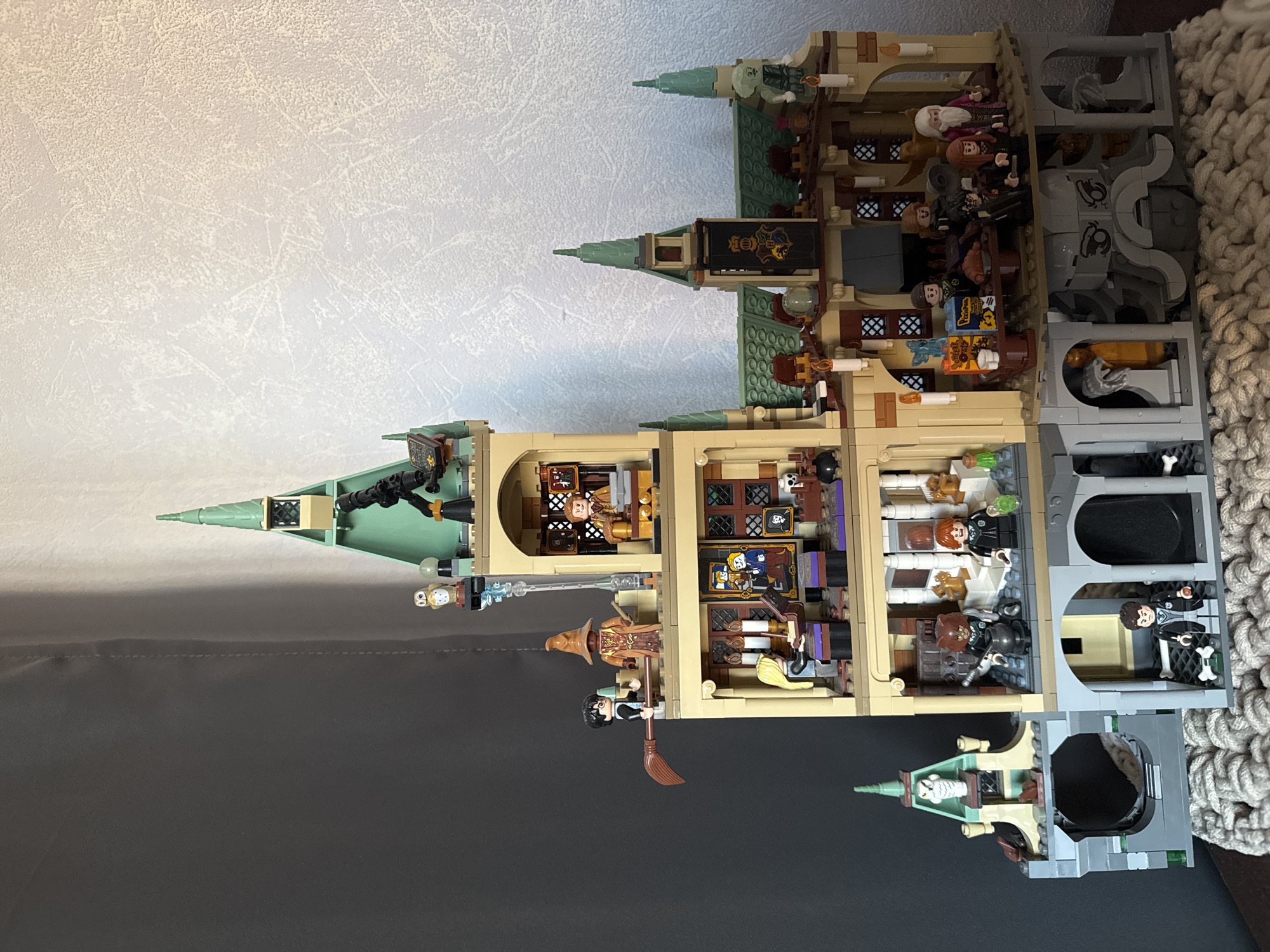 The image shows a Lego model of Hogwarts castle. The castle is built in sections to reveal the interior rooms, populated by Lego minifigures. There are different levels and towers visible. The castle is placed on a surface that transitions from a dark gray to a light gray background. A textured, possibly knitted, item is partially visible on the right side of the image. The image provides no information about the city of Moscow or Russia.