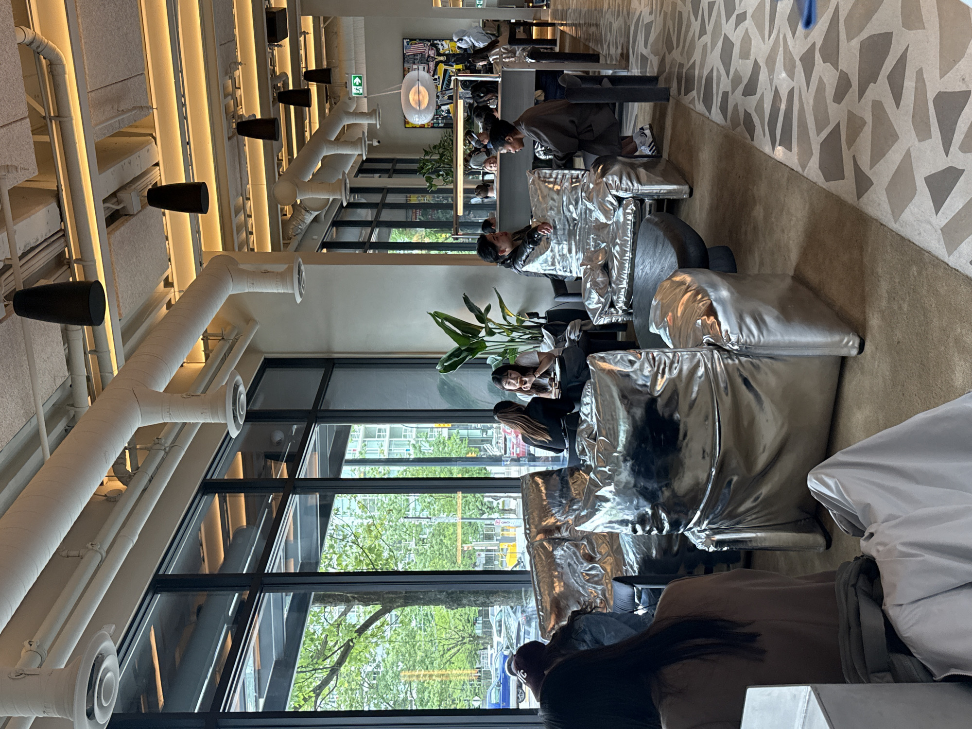 The image captures the interior of a cafe or lounge space in Vancouver, Canada. Large windows offer a view of a street with trees and city buildings, revealing the location. Several individuals are seated in modern, silver-colored beanbag chairs. Some are engrossed in their devices, while others appear to be engaged in conversation. The room's decor features exposed white ductwork and industrial-style lighting fixtures on the ceiling. An abstract art piece hangs on the wall at the back. The flooring has a geometric pattern. The overall ambiance suggests a contemporary and urban setting.