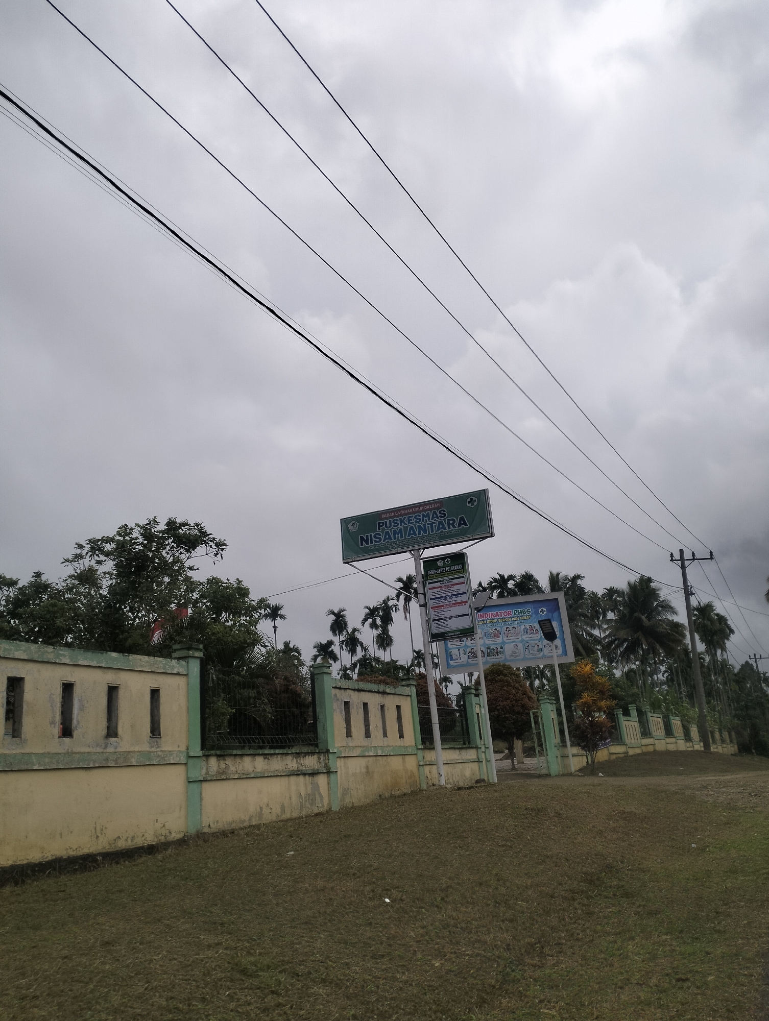 The image shows the entrance to a public health center, "Puskesmas Samantara," in Aluepapeun, Indonesia. A light yellow wall with green trim runs along the foreground, separating a grassy area from the facility. Several power lines are visible crisscrossing the overcast sky. Behind the wall, a gate leads to the Puskesmas. Two large billboards are displayed near the entrance, one with "Puskesmas Samantara" and another featuring information about healthcare services. Palm trees and other foliage are visible behind the building. The sky is heavily clouded, suggesting an overcast or potentially rainy day. No people are visible in the scene.