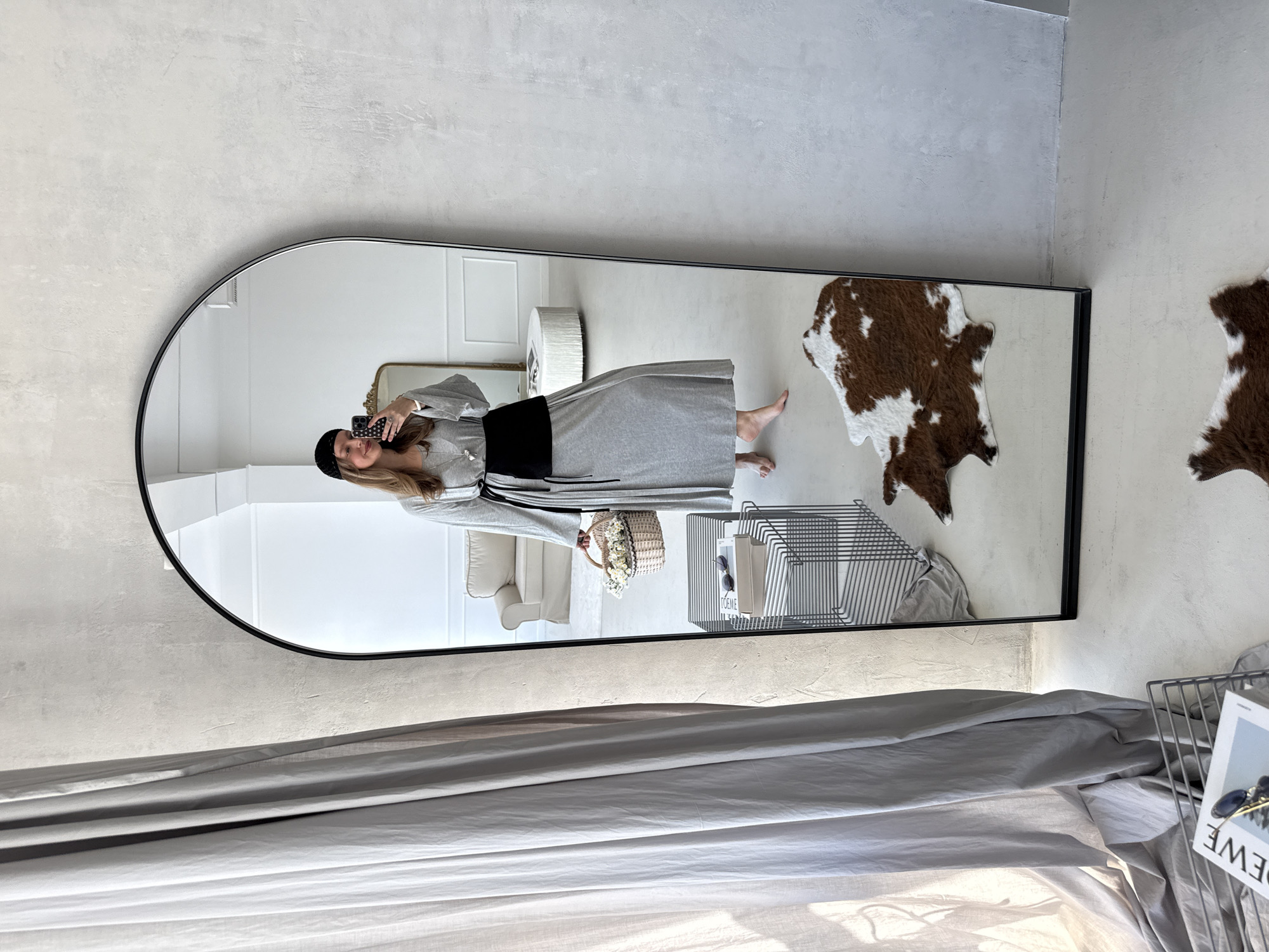 A woman is reflected in a large, arched mirror, taking a selfie. She is wearing a grey long-sleeved dress cinched with a wide black belt, a black beret, and a necklace. She holds a small wicker basket filled with white flowers. Her bare feet are visible. The mirror is positioned against a textured, light grey wall. To the left of the mirror, a grey curtain hangs. In the reflection, a beige sofa and a round textured ottoman are visible in the background. On the floor in front of the mirror is a cowhide rug. To the right of the mirror, a stack of books and sunglasses rests on a black wire shelf. In the foreground, a white magazine with the word "LOEWE" is visible on a black wire shelf. The scene appears to be an indoor setting, likely a home.