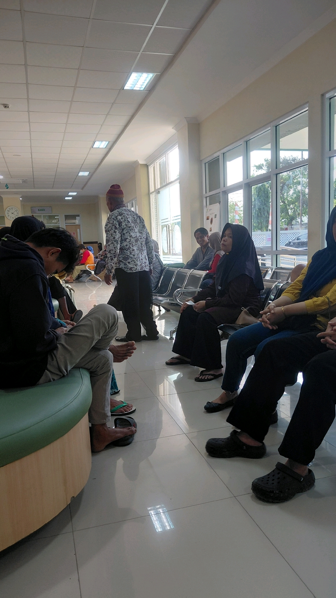 A medium shot captures a waiting area in what appears to be a clinic or hospital in Rejasari, Indonesia. The room is brightly lit by fluorescent ceiling lights, reflecting off the polished tiled floor. A row of metal chairs lines the wall, occupied by several individuals.
In the foreground to the left, a young man wearing a black hoodie and light gray pants is seated on a green bench, looking down at his lap. Next to him, a woman in a blue headscarf and traditional clothing sits on the floor with her legs crossed. To her right, a woman in a dark hijab and a maroon long-sleeved shirt is seated in one of the chairs, holding a white paper. To her right, another woman, wearing a yellow long-sleeved shirt and a dark blue hijab, is also seated, with her hands clasped.
Further back, a man in a patterned shirt and dark pants stands facing away from the camera, towards a group of people seated in the chairs. Another man is seated in the chairs to the right, smiling.
The environment is functional and clean, with large windows along the right wall offering a view of trees and buildings outside. A clock is visible on the wall in the background, along with a sign that reads "KASIR" (cashier). The overall atmosphere suggests a typical waiting area scenario.