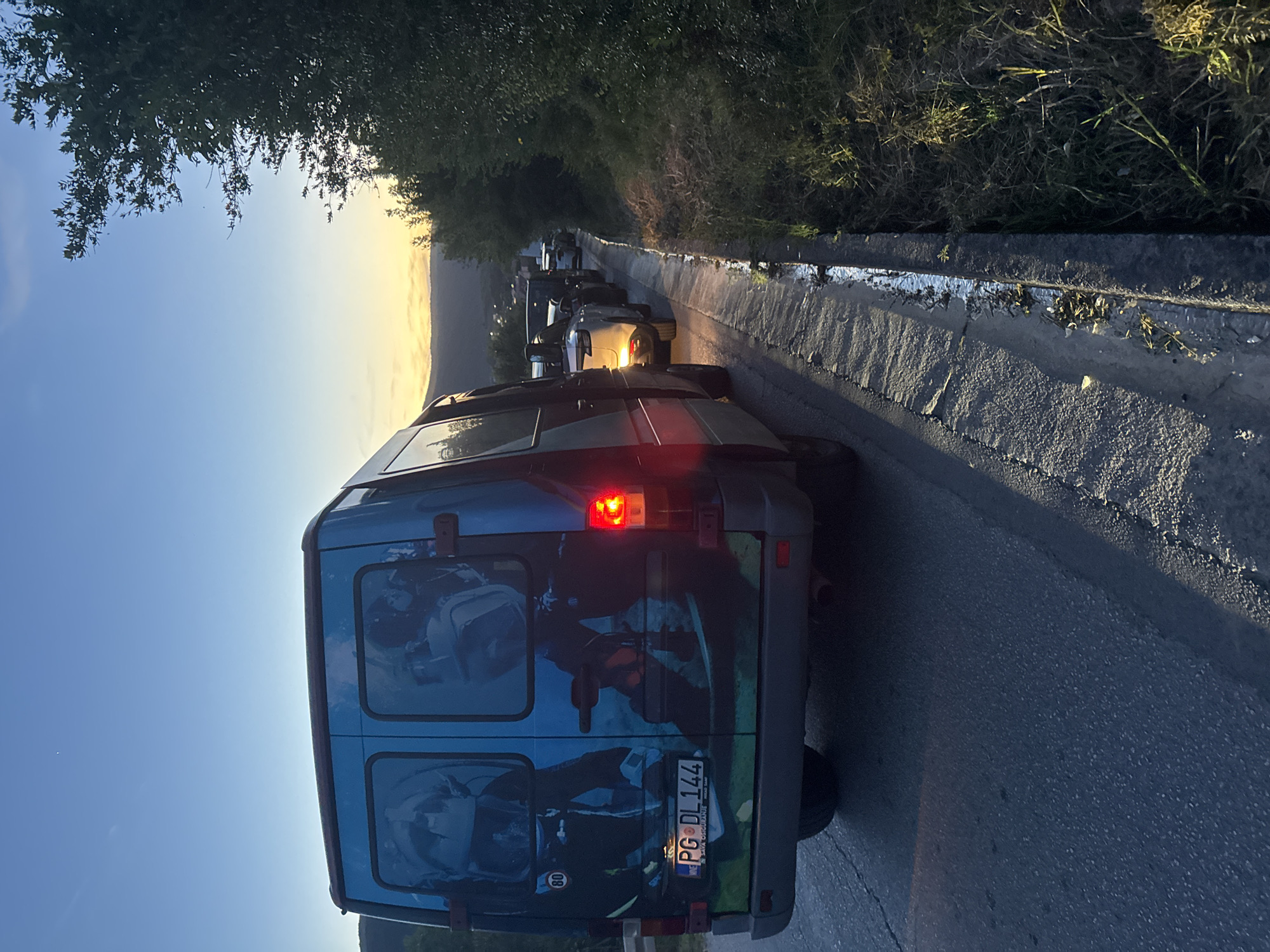 An outdoor eye-level shot captures a line of vehicles on a road, illuminated by the low sun at either sunrise or sunset. In the immediate foreground, facing away from the viewer, is a blue van with its right brake light illuminated, suggesting it is stopped or moving slowly. The van features custom graphics on its rear, depicting blue and gray adventure-themed imagery, possibly figures with backpacks or engaged in outdoor activities. A white circular sticker with "80" is visible on the left rear, and its license plate reads "PG DL 144," with smaller text "SVA OSIGURANJE" above the main numbers, indicating a registration from Podgorica, Montenegro.
Beyond the blue van, a queue of other cars stretches into the distance along a winding road, their headlights or reflections gleaming in the golden light. The road is paved, with a concrete curb and an overgrown, grassy shoulder on the right side. Lush green trees and foliage line both sides of the road, particularly prominent in the upper left of the frame. The sky transitions from a deep blue at the top to a bright, golden yellow and orange near the horizon, where distant hills or mountains are silhouetted against the bright light. The overall scene suggests a moment of pause or slow movement on a scenic route during the early morning or late evening.