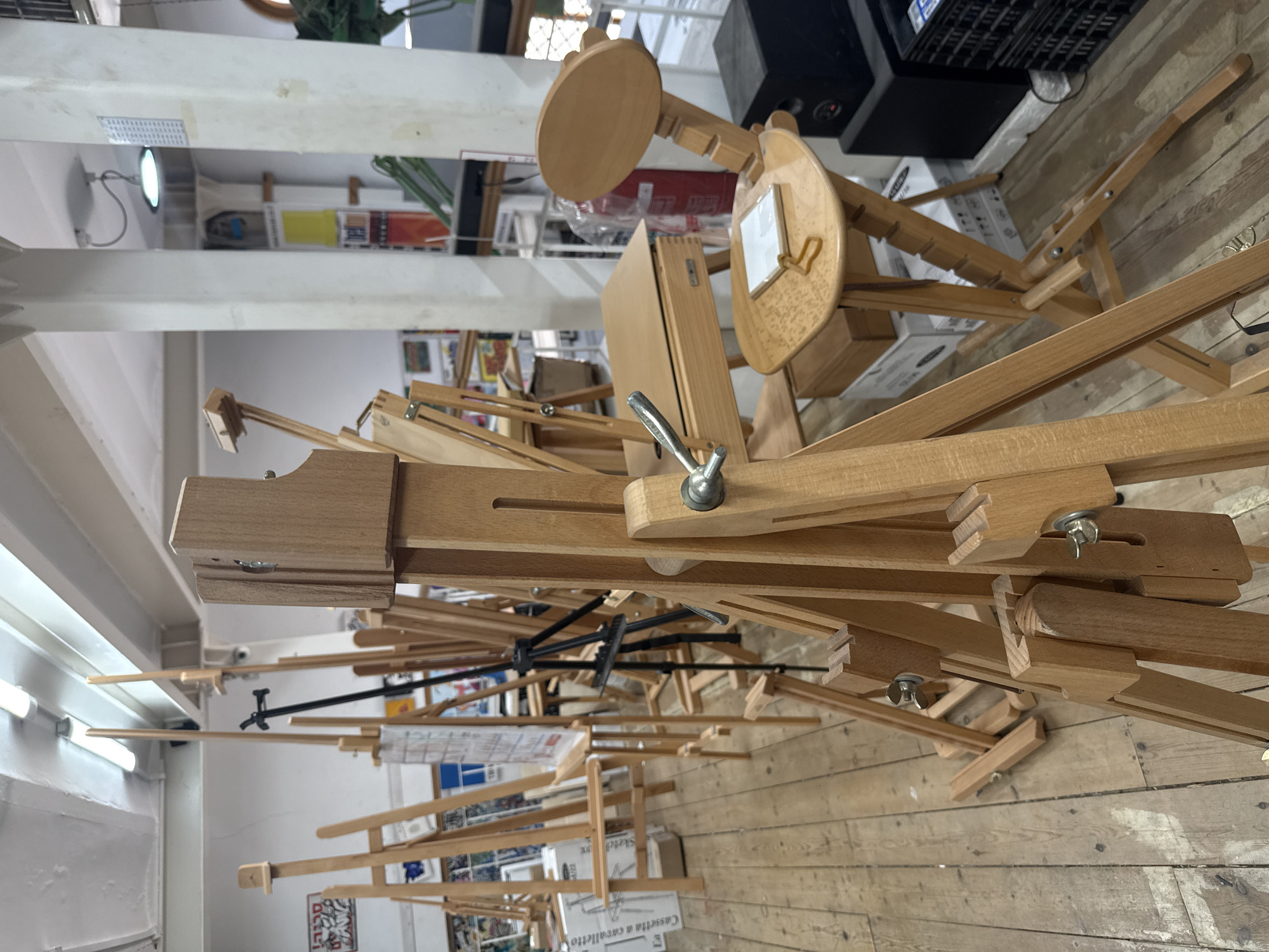 The image displays an indoor space, likely an art supply store or studio, filled with numerous wooden easels. The perspective looks slightly down into a densely packed area.
The primary subjects are various wooden easels, predominantly made of light-colored wood. They are of different designs, some fully assembled and standing tall, while others appear folded or stacked, showcasing their adjustable features like slotted wooden tracks, metal handles, and bolts. One easel in the upper portion of the frame features a small, round wooden table or palette, and another has a small white canvas or board affixed to its painting rest.
The setting includes a wooden plank floor and white walls or structural beams. Artificial illumination is provided by fluorescent strip lights visible on the ceiling or upper walls. In the background, shelves hold various items, and a black speaker or electronic device is visible.
No people are present, and there is no specific activity depicted, suggesting a retail display, storage area, or a calm moment within a creative environment.
Visible text on a white box on the floor reads "Cassetta a cavalletto" (Italian for "Easel box") and partially "Sketch". The overall atmosphere is functional and dedicated to artistic endeavors.
