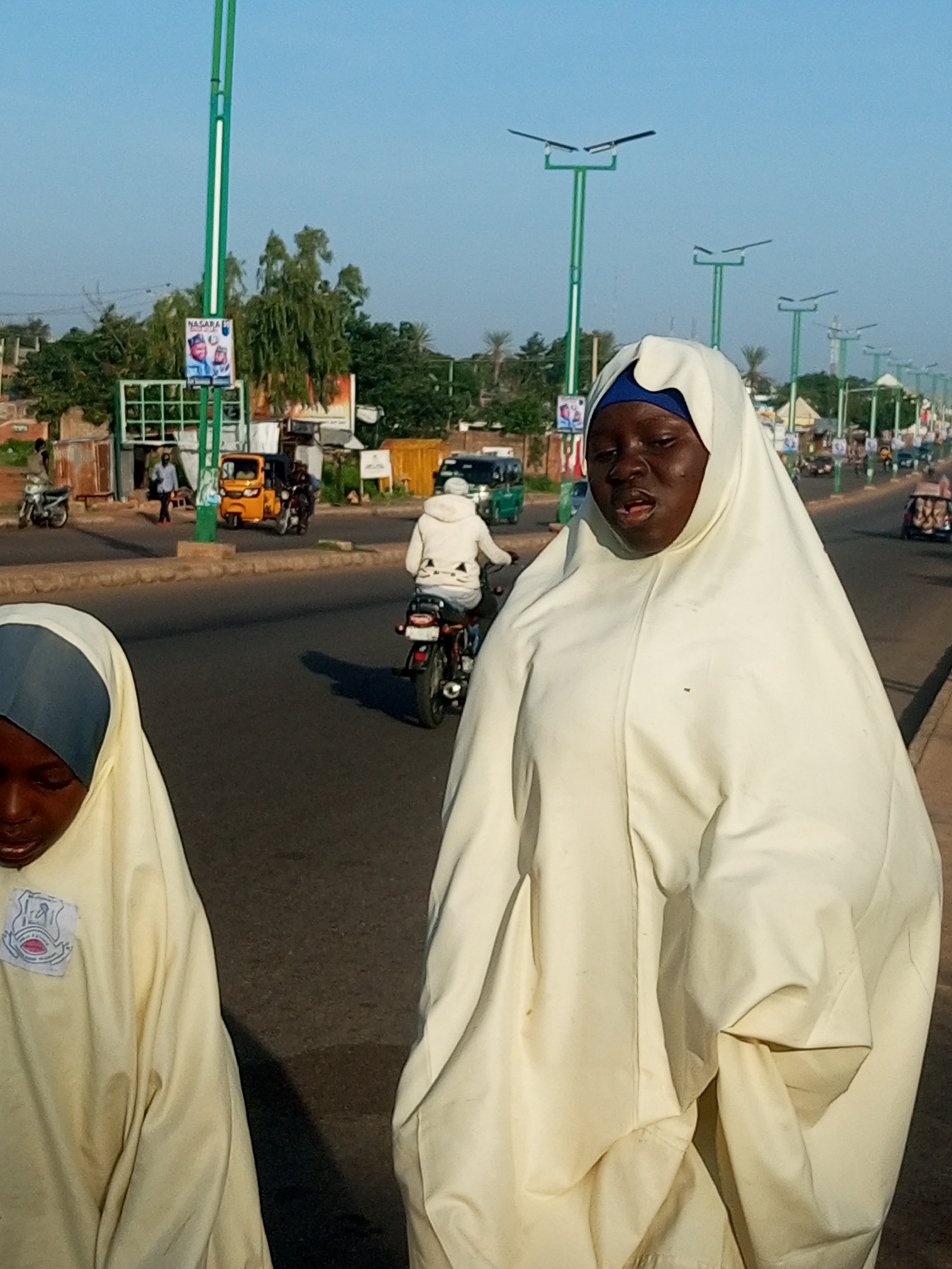 In Gombe, Nigeria, a street scene unfolds with two young girls dressed in light-colored Islamic veils in the foreground. The girl on the left is looking down, while the girl on the right is looking ahead with her mouth slightly open. Behind them, traffic moves along a paved road lined with lampposts. A yellow auto-rickshaw is visible, along with motorcycles and cars. Buildings and trees form the backdrop, under a clear blue sky. A poster with two faces is attached to one of the lampposts.