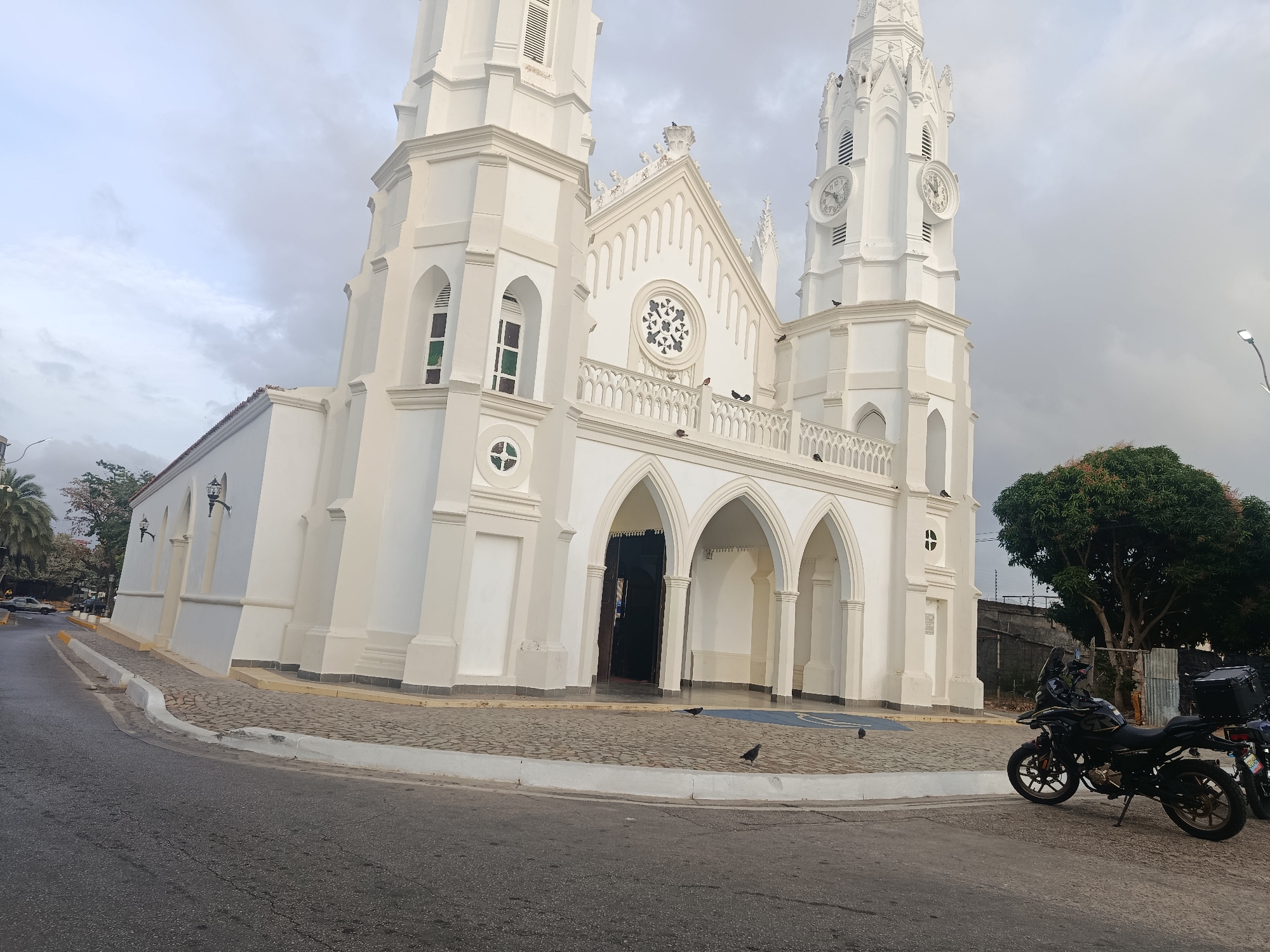 The image shows a white, ornate church with two clock towers, situated in Juan Griego, Venezuela. The church has arched entrances and a rose window. A street runs along the front of the church, and two motorcycles are parked on the street near the church. Several birds are visible on the ground near the church entrance. A tree is next to the church. The sky is cloudy.