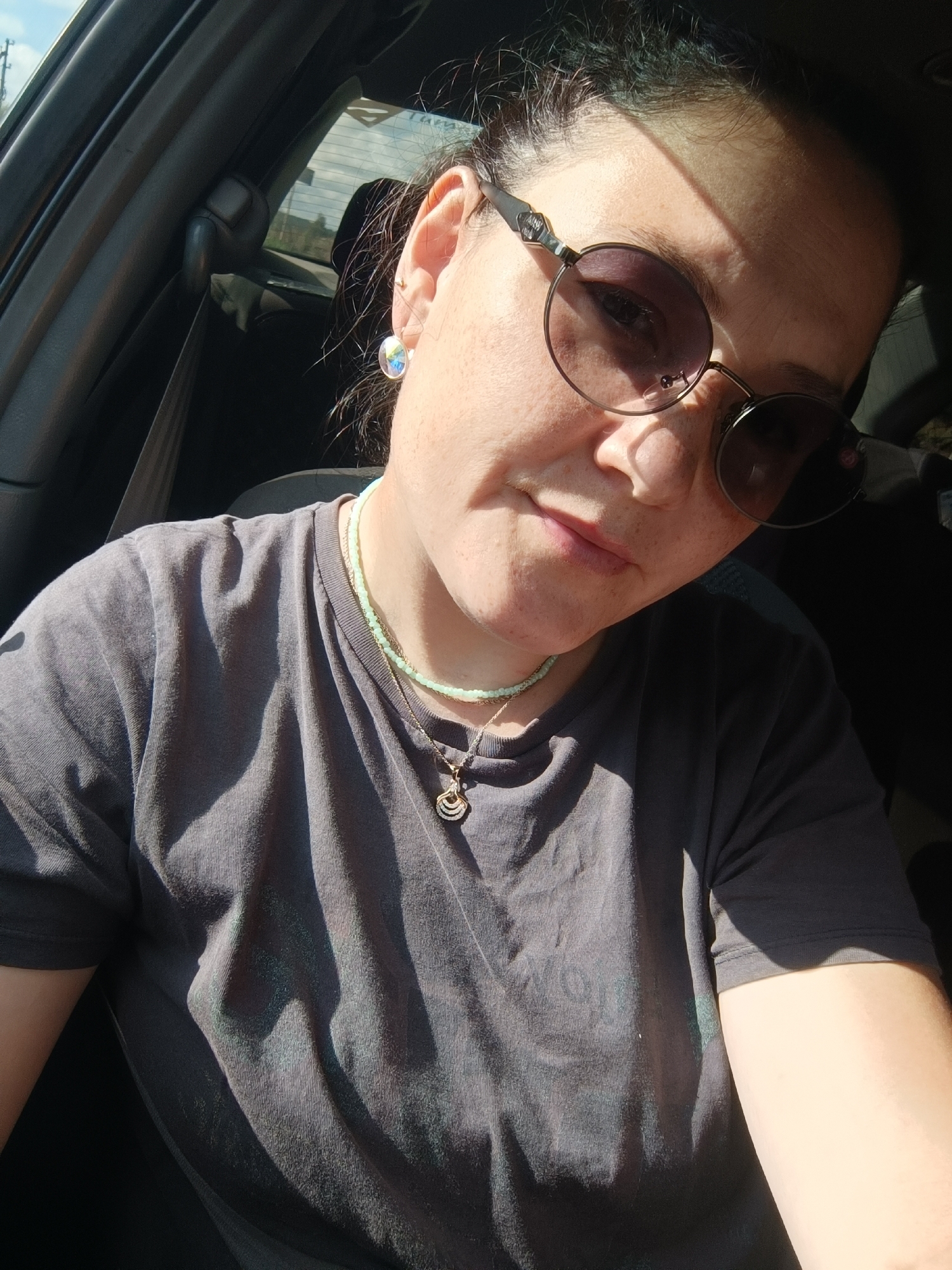 A medium shot captures a woman from the chest up, seated in the driver's seat of a vehicle. She is wearing a dark gray t-shirt, a light green beaded necklace, and a necklace with a curved pendant. Round, dark-tinted sunglasses obscure her eyes, and a faint smile plays on her lips. The lighting suggests it is daytime, with sunlight casting shadows across her face and clothing. The interior of the car is visible, including a seatbelt. The location is identified as Foki, Russia.