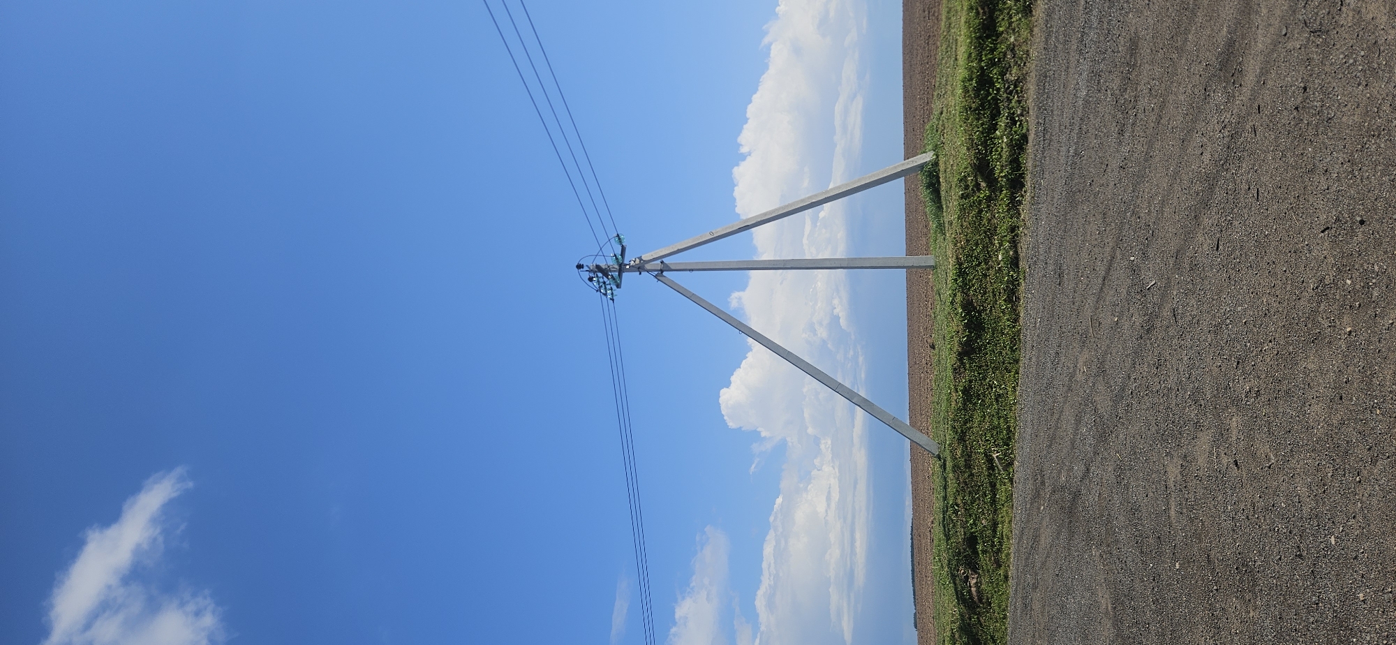 A concrete utility pole with a triangular base stands in a field under a blue sky with scattered white clouds. Three black power lines extend from the top of the pole. The field is covered in brown soil and green grass. A gravel road is visible in the foreground.