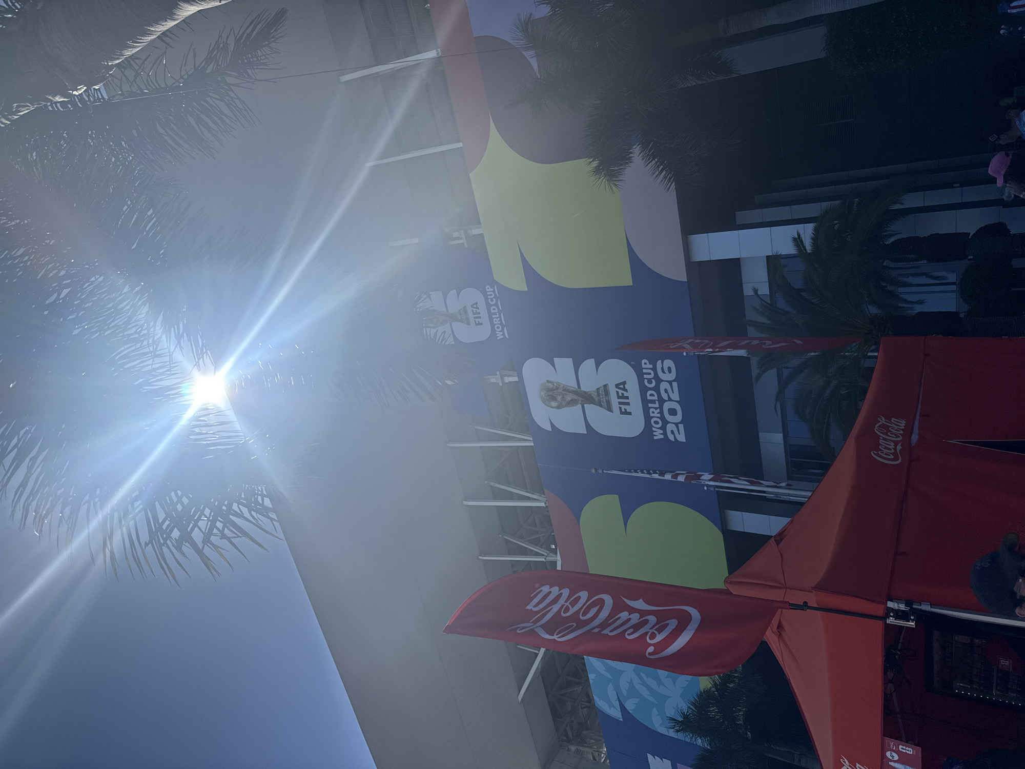 In Miami Gardens, Florida, a bright sun shines over a stadium decorated with a banner for the FIFA World Cup 2026. A red Coca-Cola flag and a red tent are in the foreground. The banner features the World Cup trophy and the number "26". Palm trees are visible throughout the scene.