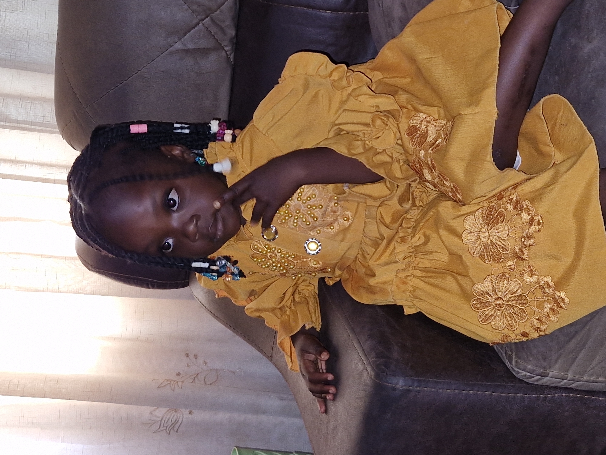 A young girl with braided hair adorned with beads and pink and blue accents sits on a brown leather couch. She is wearing a mustard-yellow dress with ruffled sleeves and a ruffled hem. The dress is decorated with gold beads and embroidery. Her right hand is resting on the armrest of the couch, and her left hand is near her mouth. She is looking to her left. The background consists of a sheer, light-colored curtain with subtle floral patterns. The setting appears to be an indoor residential environment, consistent with an apartment or home in Jos, Nigeria.
