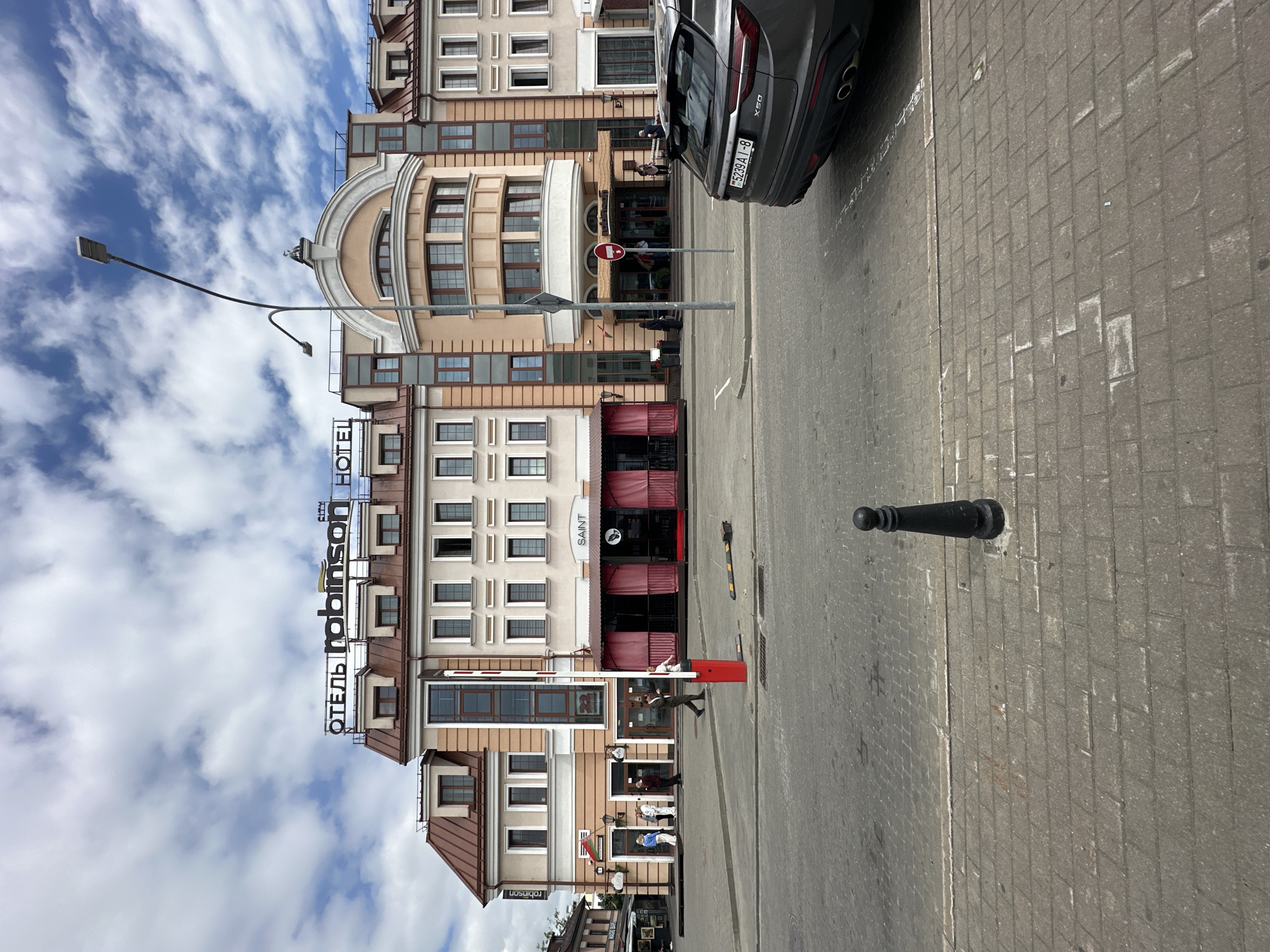 The image depicts the Robinson Hotel in Minsk, Belarus. The hotel building features a prominent sign with the hotel's name in both Cyrillic and Latin script. The architecture is multi-story with a mix of light-colored facade and darker window frames, topped with a mansard roof. In front of the hotel, a red and white barrier arm is positioned on the sidewalk, likely for traffic control. Several people are visible in front of the hotel entrances, some walking and others standing. A dark-colored SUV is parked on the right side of the image, with its rear facing the viewer. The foreground shows a paved street and sidewalk with cobblestone patterns. The sky above is cloudy, with patches of blue visible.