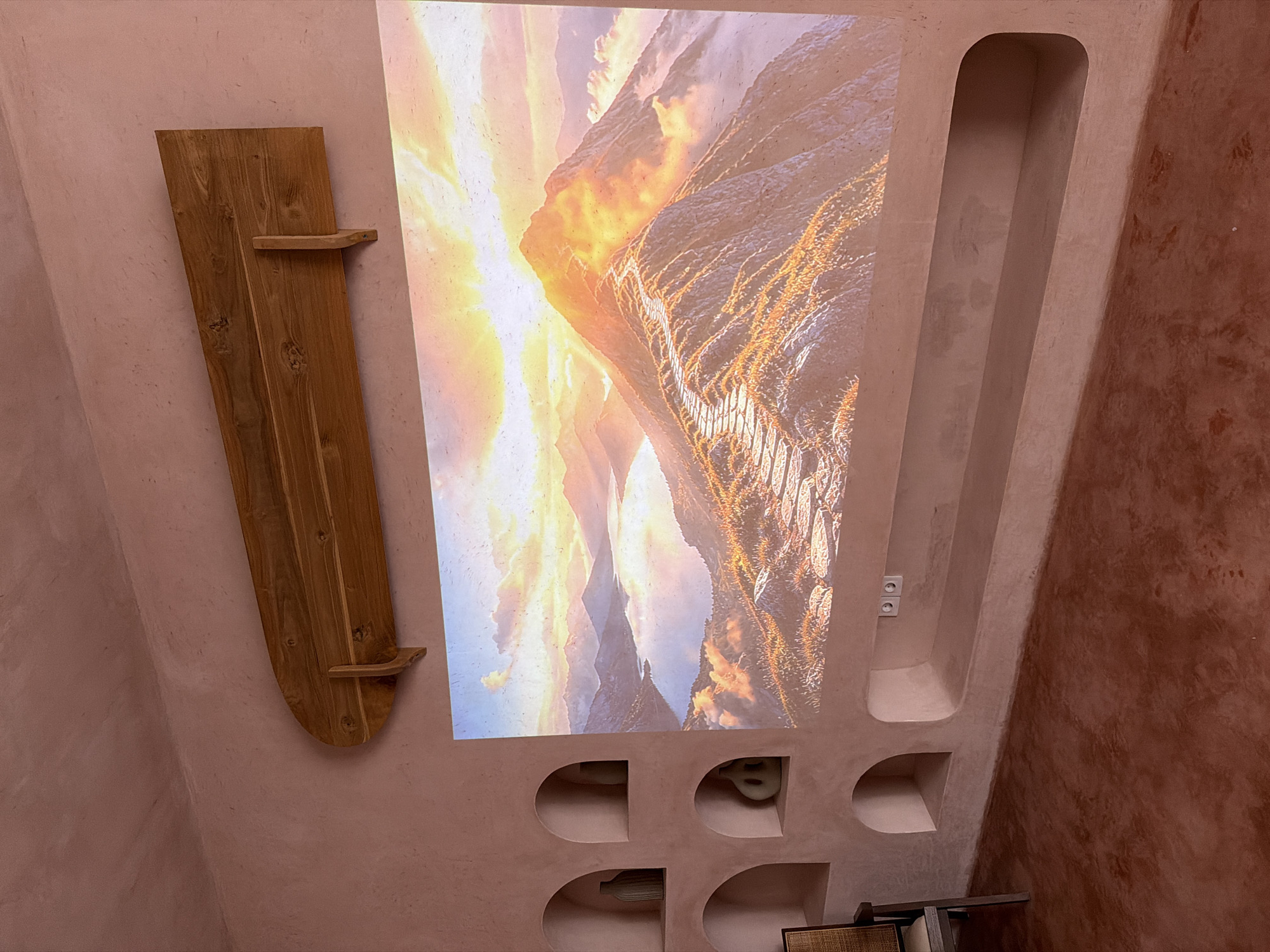 The image captures an interior scene, possibly in North Kuta, Indonesia, characterized by its unique wall design and decorative elements. The walls are predominantly a textured, reddish-pink hue, with a section featuring a deeper shade of reddish-brown. On the left, a wooden decorative piece, resembling a plank with two small protruding shelves, is mounted.
In the center of the wall, a bright image is projected, depicting a landscape with mountains and a sky featuring a sunset or sunrise. The projection is contained within a recessed, angular frame that mimics the architectural style of the wall. The frame extends downwards to incorporate a series of semicircular cubbies, each appearing empty, except for one that may contain a small object. Above the cubbies, two electrical outlets are visible within the frame.
To the right of the projected image, a vertical, recessed channel extends from the top of the wall, tapering slightly downwards. The wall’s angular design is complemented by the textures and colors, creating a somewhat rustic and modern aesthetic. Part of a chair or another piece of furniture with a woven seat is visible at the bottom of the frame.