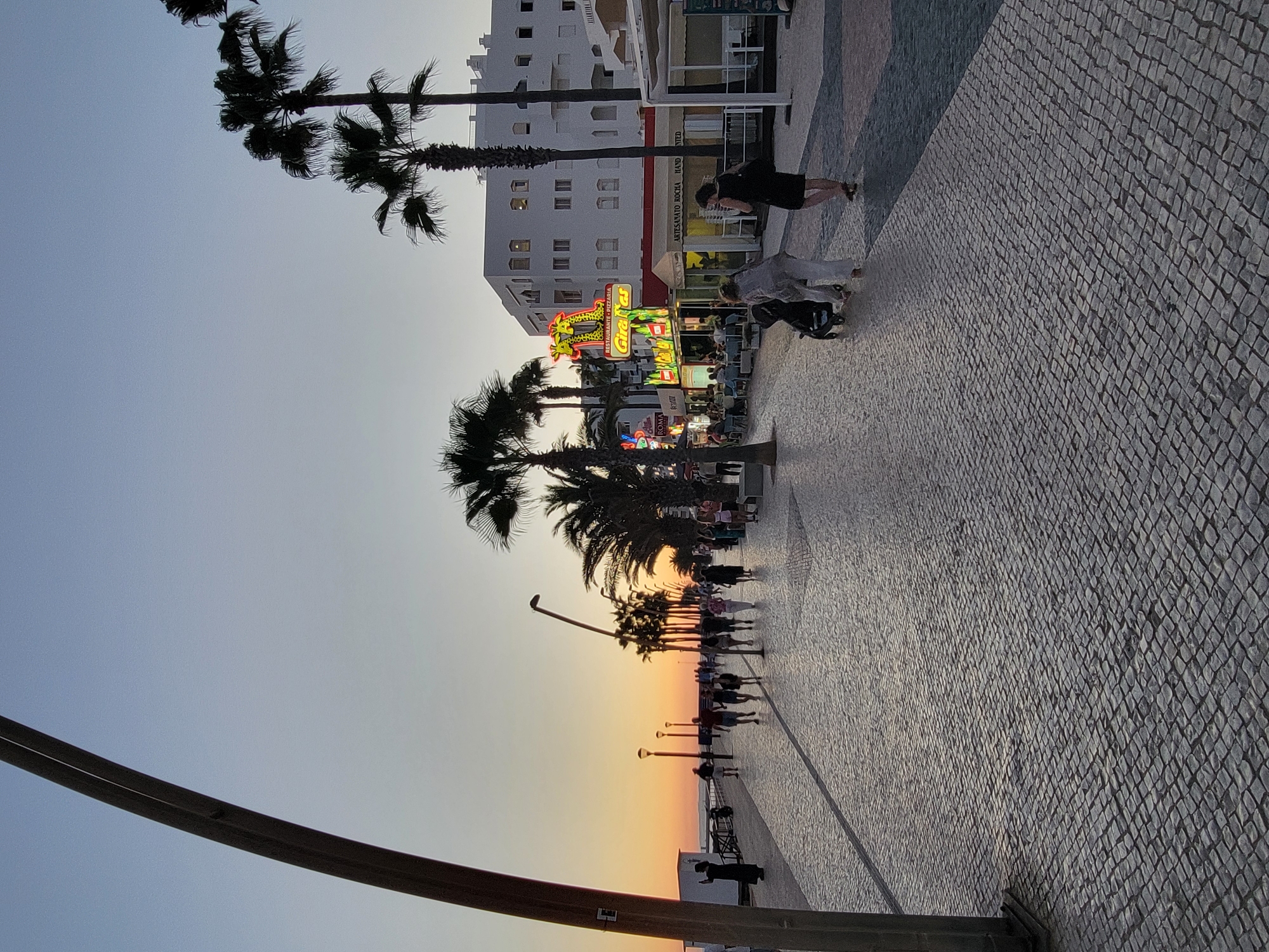 This image depicts a sunset scene on a promenade in Quarteira, Portugal. The promenade is paved with cobblestones and lined with palm trees. A white building with multiple windows stands in the background, featuring several businesses with neon signage, including one for "Restaurante Pizzeria Girafas." Several people are walking along the promenade, some are seated at outdoor tables in front of the restaurants. A lamppost is visible on the left side of the image, and a dark pole runs vertically along the left edge. The sky transitions from orange at the horizon to a pale blue overhead. Two women, one pushing a stroller, are walking away from the camera in the foreground.