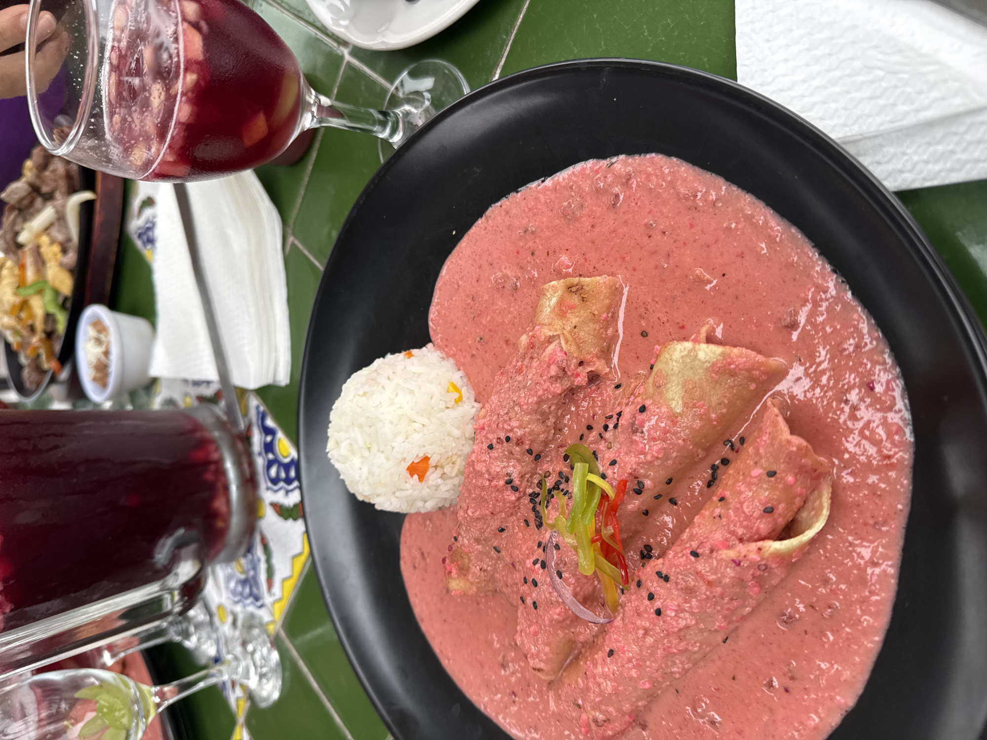 The image shows a close-up of a dish of enchiladas served with rice, accompanied by a glass of sangria on a table with a green tiled surface. The enchiladas are covered in a pink sauce and sprinkled with black sesame seeds. The rice is topped with small pieces of orange and yellow pepper. In the background, a pitcher of sangria and another glass of the drink are visible, along with a plate of what appears to be fajitas. The scene suggests a dining experience at a restaurant, potentially in Taxco, Mexico, given the mention of the city. The lighting is bright, indicating it is daytime.