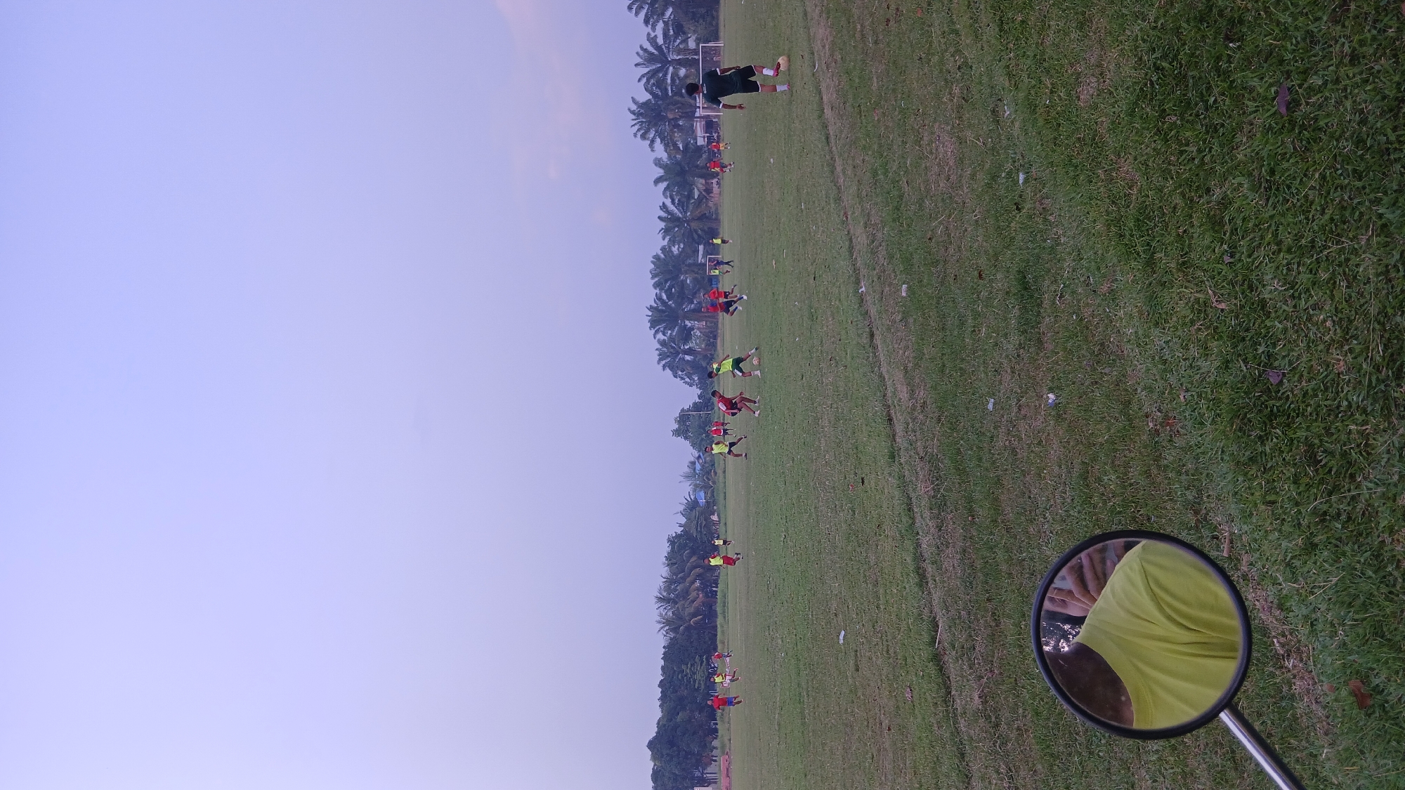 The image depicts an outdoor soccer game on a grassy field, likely in Klumpang Kebun, Indonesia, under a pale sky. Several individuals are actively engaged in playing soccer, wearing various colored jerseys, predominantly red and yellow. One individual, dressed in a dark jersey with white trim and black shorts, stands with a soccer ball at his feet in the foreground. The field is bordered by a line of palm trees and other vegetation, suggesting a tropical or subtropical climate. In the lower left quadrant of the image, a motorcycle's rearview mirror reflects the shoulder and neck of a person wearing a bright yellow shirt. The overall scene captures a moment of athletic activity in a rural or semi-rural setting.