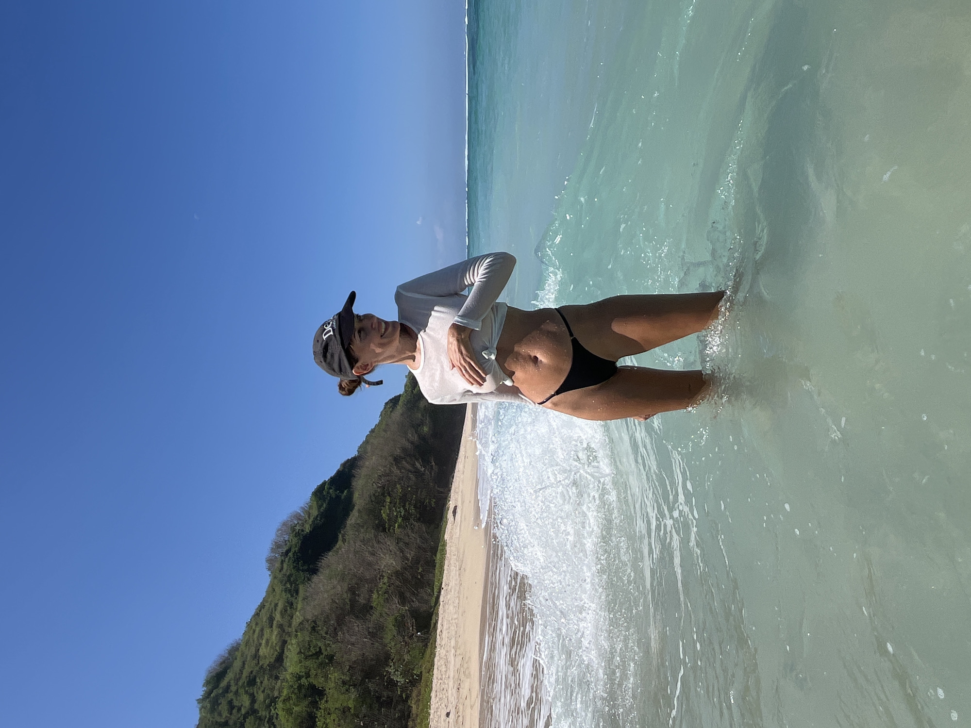 In Kuta Selatan, Indonesia, a woman stands in the shallow, turquoise waters of a beach. She is wearing a black baseball cap, a long-sleeved white cropped shirt, and black bikini bottoms. Her hands are resting on her chest, slightly lifting the shirt. The ocean waves are breaking around her legs, creating a foamy effect. In the background, a sandy beach meets a lush, green hillside under a clear blue sky.