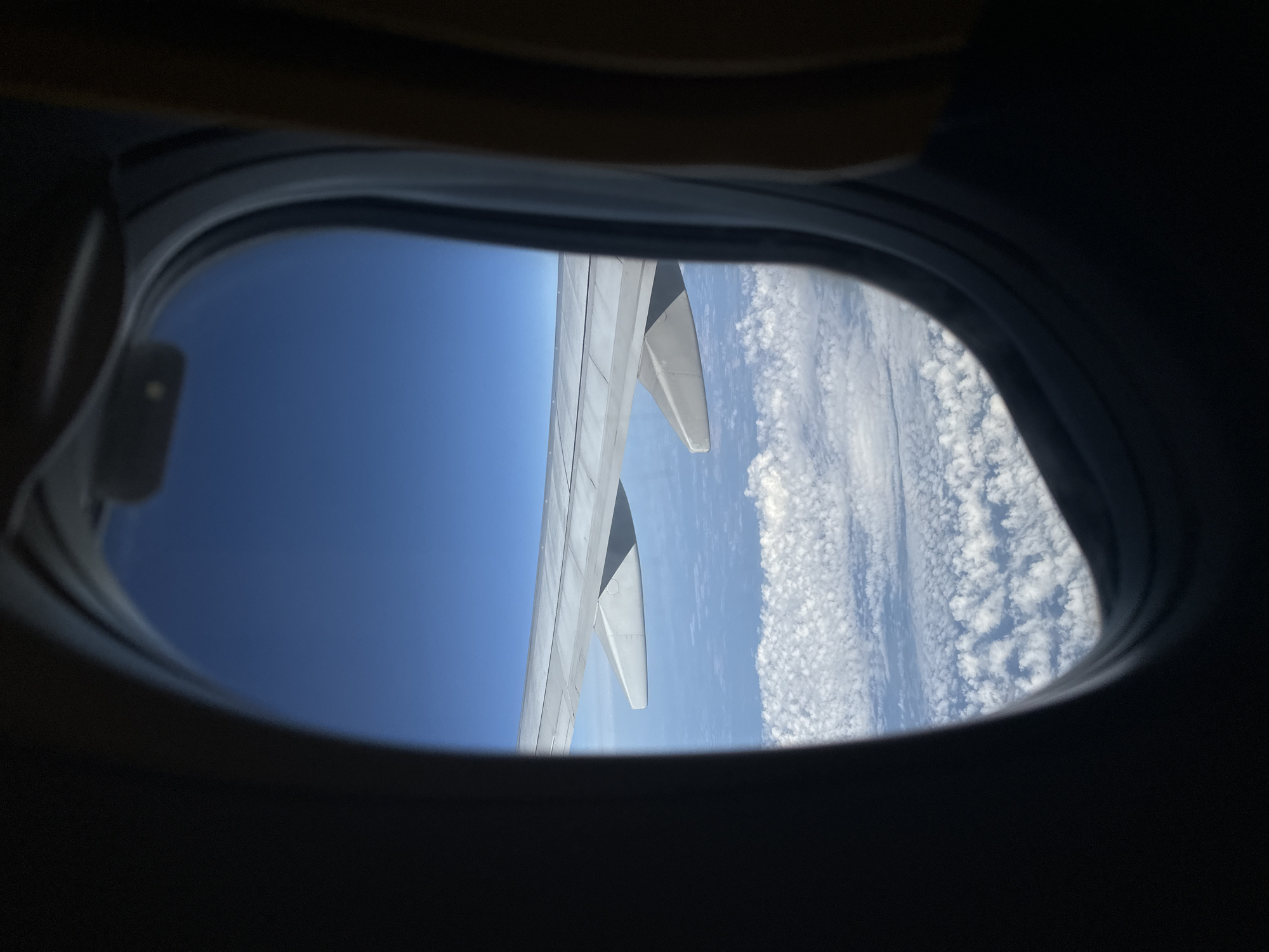 The image is an interior view from an airplane window. A portion of the airplane wing extends into the frame from the left. The wing is light gray and features a downward-angled flap near its end. Below the wing, a vast expanse of white, puffy clouds stretches across the scene. The sky above is a clear, deep blue. The window frame, which is dark and oval-shaped, partially obscures the view. The right side of the frame shows a glimpse of a seat, upholstered in a yellowish-brown fabric. The perspective suggests the airplane is in flight, high above the clouds. The location in the city of Nagyhajmás, Hungary is not discernible from this image.