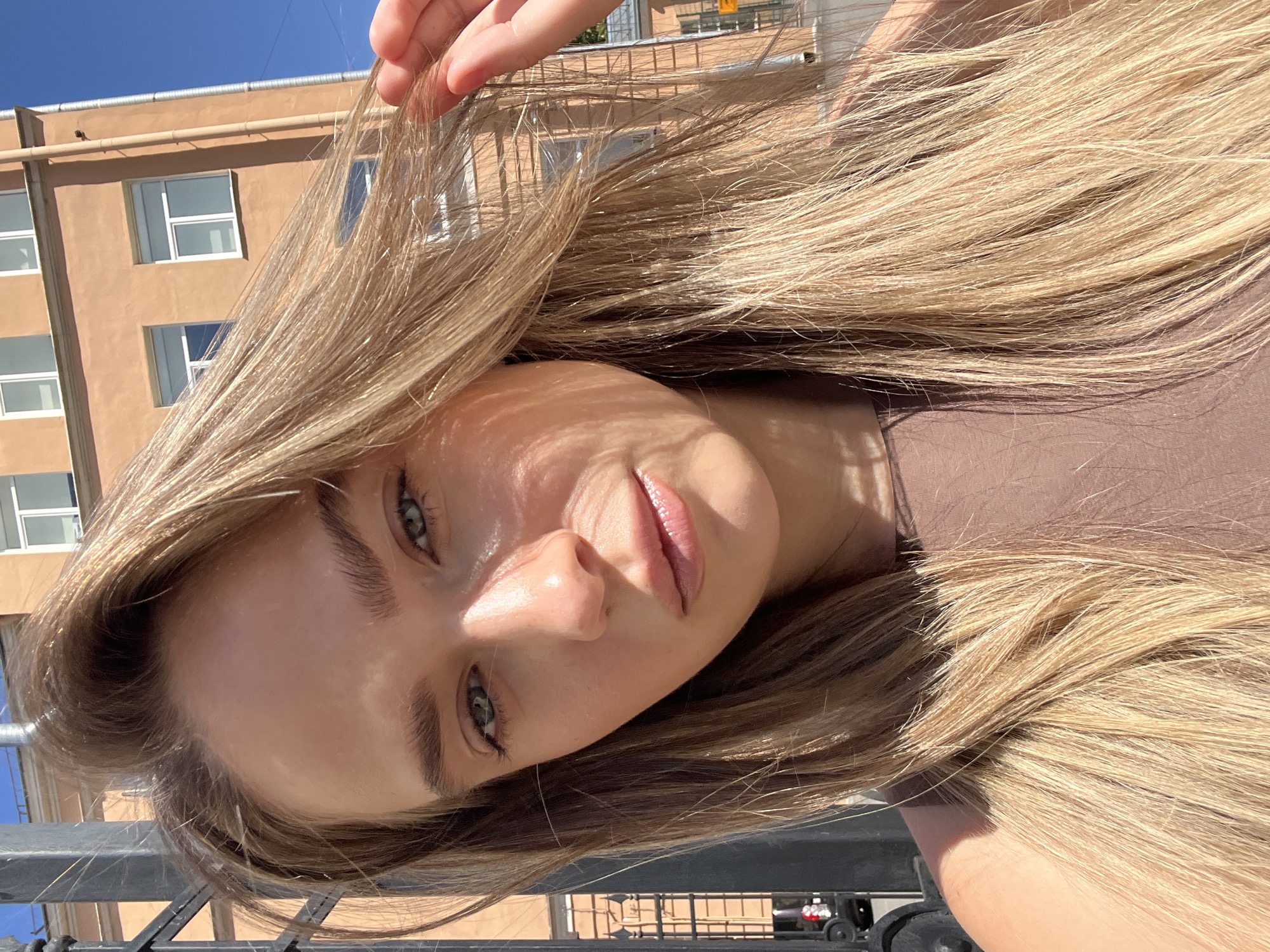 A close-up selfie of a young woman with long blonde hair and blue eyes. She is outdoors on a sunny day, with a tan building and a clear blue sky in the background. A black metal fence is visible on the left side of the image. The woman is wearing a brown top. The lighting suggests a midday sun. The architecture of the building indicates it is likely located in Saint Petersburg, Russia, possibly in an older, residential area.