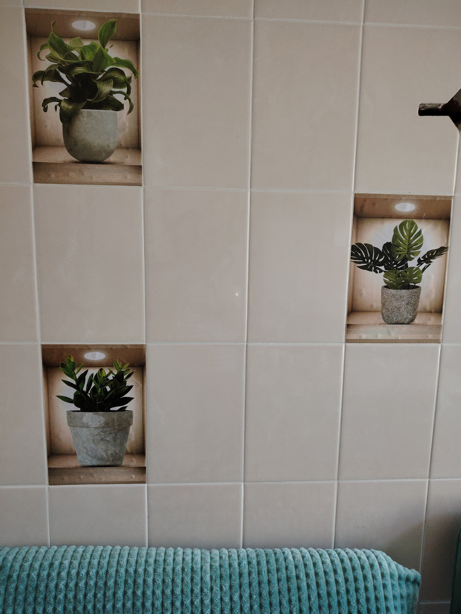 This media file displays a tiled wall with decorative decals of potted plants set into recessed niches. There are three such niches visible. The top left niche features a large, leafy green plant in a light gray pot. The middle right niche contains a plant with broad, dark green leaves, also in a speckled gray pot. The bottom left niche shows a smaller, bushy green plant in a gray pot. Each niche appears to have a light source at the top, casting a gentle glow. A teal-colored towel with a ribbed texture is draped across the bottom of the frame, obscuring the very bottom of the wall. The tiles are a neutral, light beige color and are laid in a grid pattern. In the upper right corner of the image, a dark fixture, possibly a faucet or showerhead, is partially visible. There is no text present in the image. The setting appears to be a bathroom or similar enclosed space, indicated by the tiles and towel. The lighting suggests it could be daytime, but the artificial light within the niches provides an even illumination, making a specific time of day difficult to determine.