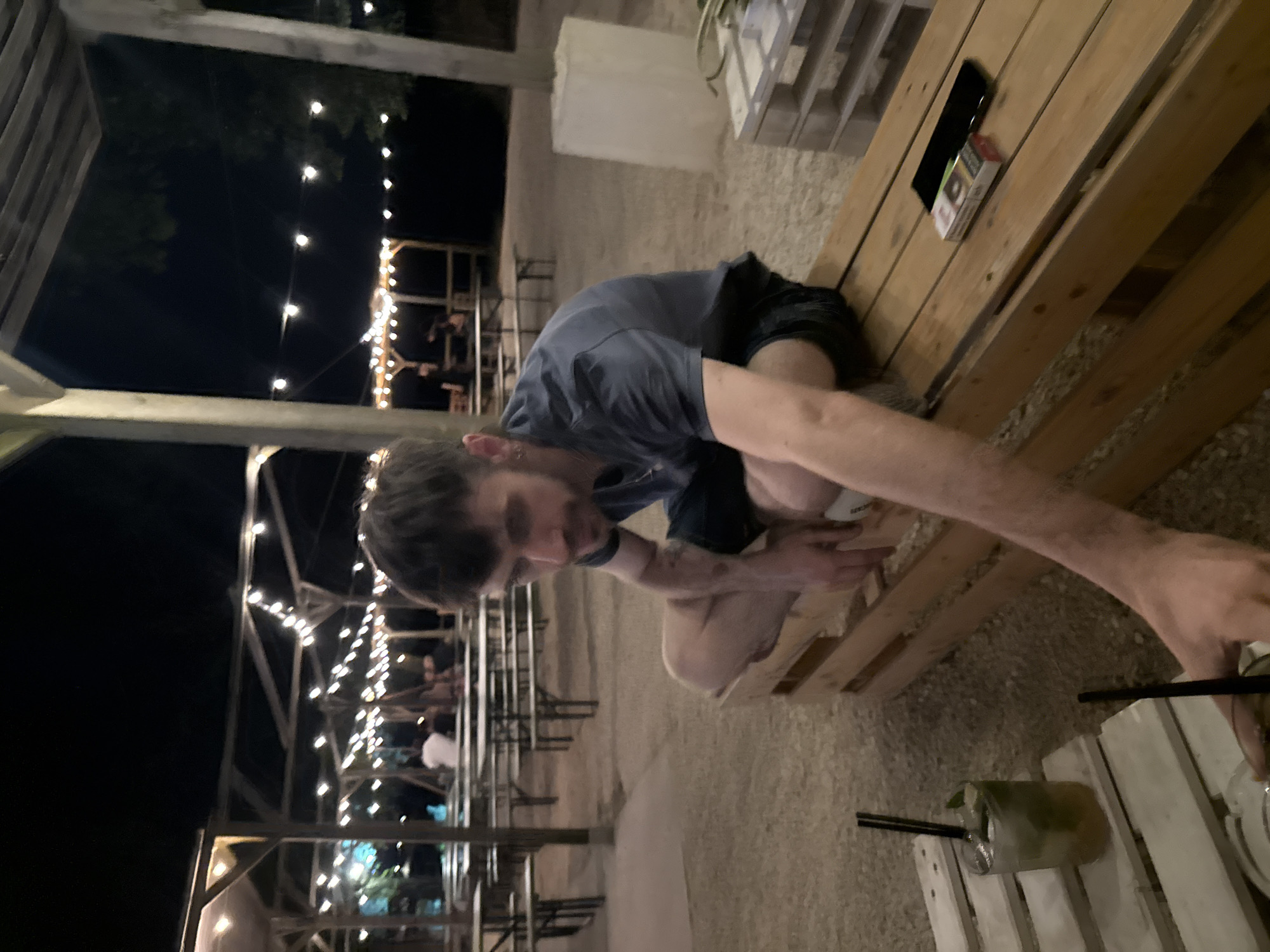 A man sits outdoors at a bar with lights in Casalmaggiore, Italy. He wears a blue t-shirt and dark shorts. He sits on a wooden pallet bench. On the bench next to him is a cell phone and a pack of cigarettes. A drink with a straw sits on a white-painted wooden table. Other patrons are seated at tables in the background under string lights.