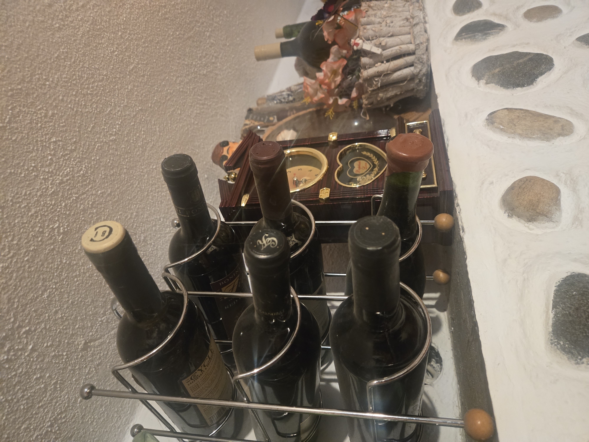 The image shows a wine rack containing six wine bottles, placed against a wall with a textured, off-white surface. Above the wine rack, there's a decorative clock in a wooden case, adorned with golden heart-shaped motifs and other ornamental details. Behind the clock, there is an arrangement featuring wooden logs and artificial flowers. To the right, a wall with a stone-like pattern is visible. The scene appears to be indoors, possibly in a cellar, wine room, or a decorative display area.