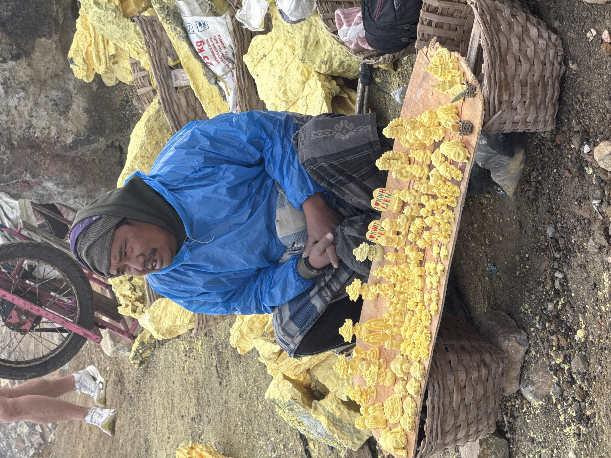 The image depicts a man in Unknown, Indonesia, sitting amid sulfur rocks. He is wearing a blue poncho and a grey head covering. A bike wheel is visible to his left. In front of him, on a wooden plank, are numerous sulfur carvings, with some bearing the words "I ❤️ KWH IJEN." Baskets flank the plank, and a sack labeled "25 kg" is nearby. The ground is covered in sulfur dust. A pair of legs is briefly visible at the bottom-left corner.