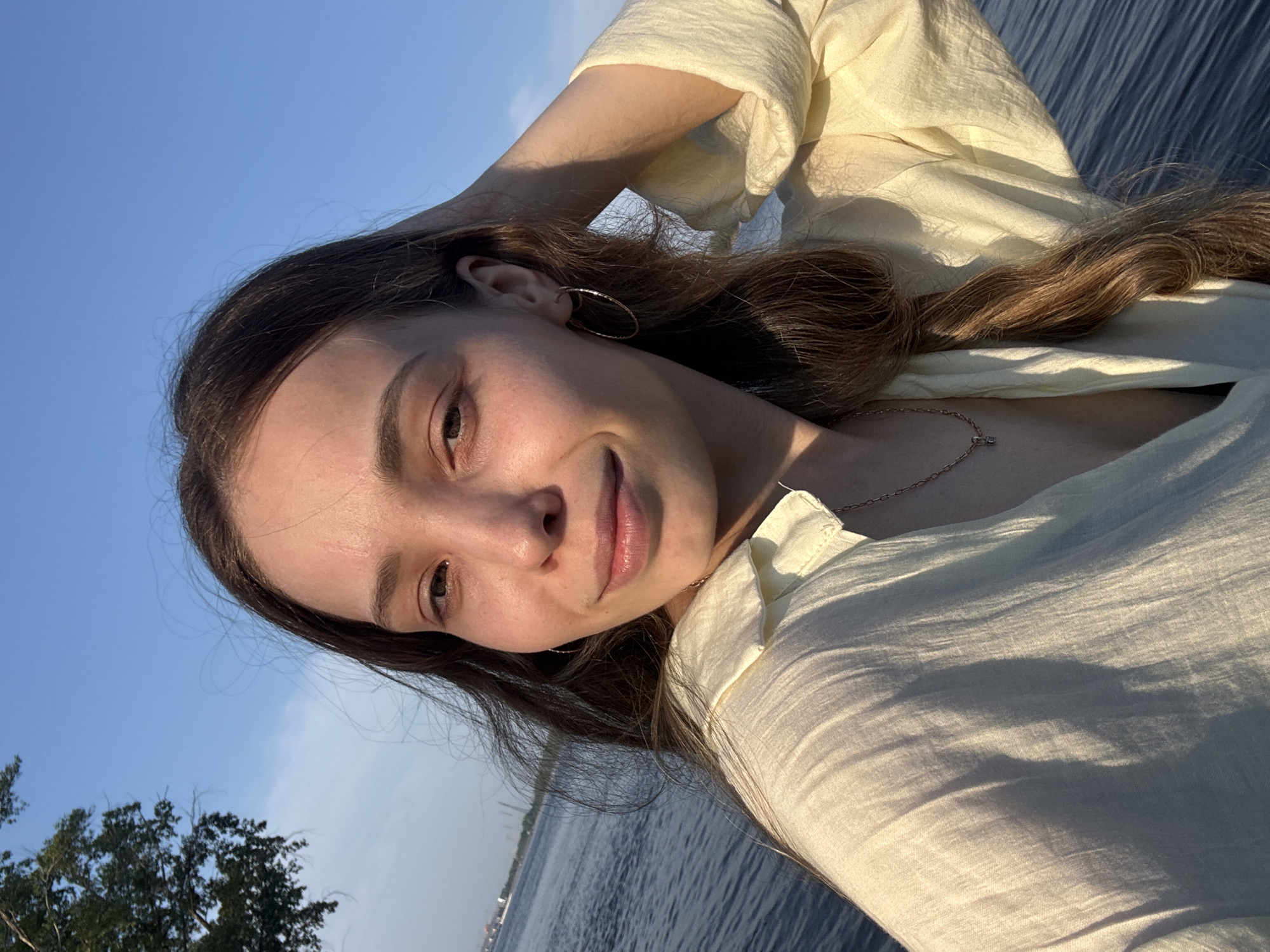A young woman with long brown hair and fair skin is pictured in a selfie taken outdoors. She is wearing a light yellow shirt and a delicate necklace. Her right hand is raised, touching her hair, and she is looking at the camera with a slight smile. The background features a body of water, a clear blue sky, and a tree on the left side. The lighting suggests it is late afternoon or early evening. The specific location is in Noviy, Russia, near the water.