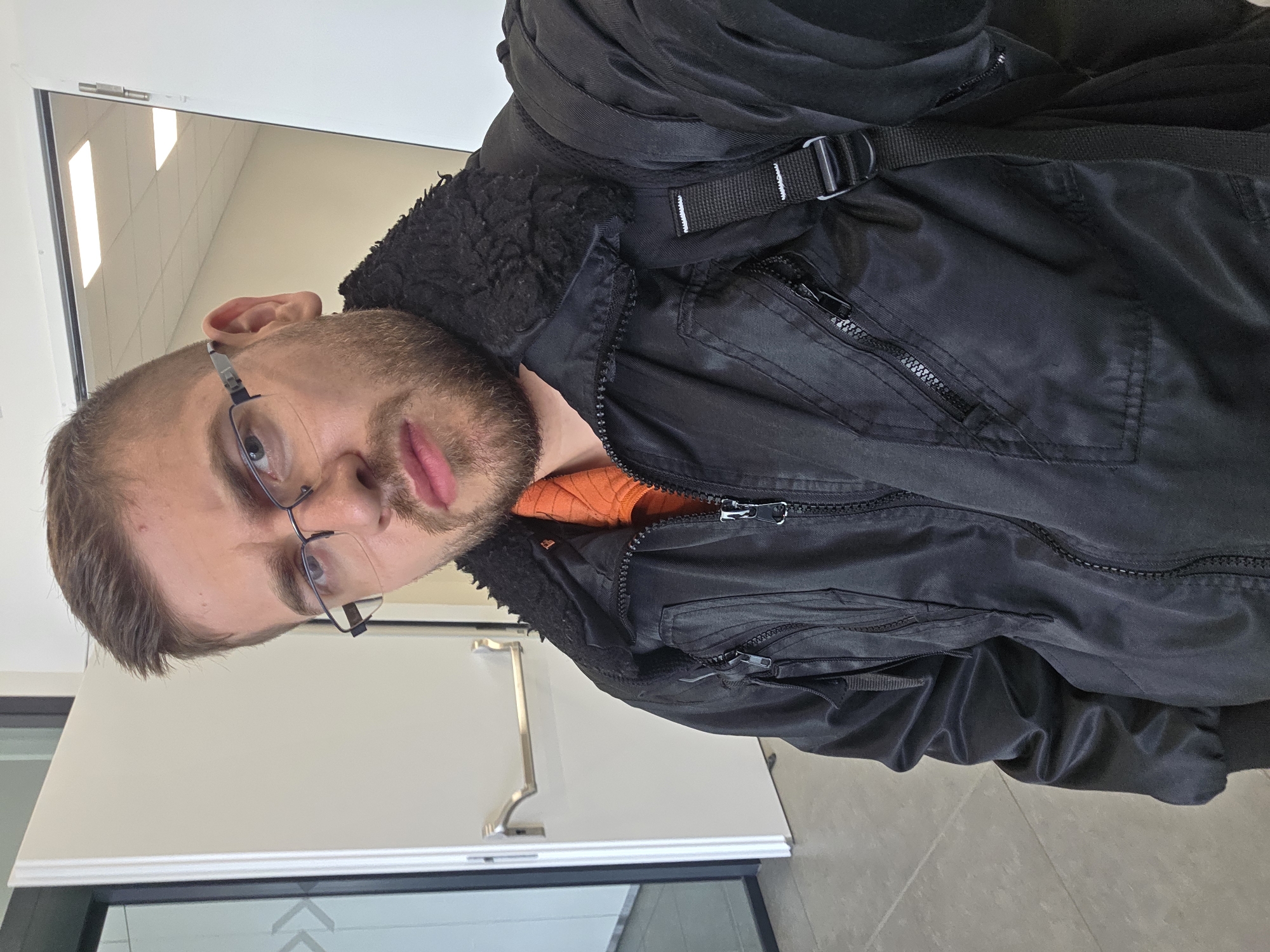 A close-up, rotated portrait captures a young to middle-aged man looking directly into the camera with a neutral expression. He has short brown hair, a neatly trimmed short beard, and wears dark, rectangular wire-rimmed glasses, which reflect the bright overhead lighting. His lips are visible and appear full.
He is dressed in a black jacket that features a prominent textured collar, likely made of faux fur or a similar material. The jacket is adorned with several zippers, indicative of pockets, and a black fabric strap with a plastic buckle is visible across his right shoulder. A bright orange garment, possibly a sweater or t-shirt, creates a striking color contrast against the dark jacket, visible underneath the lapel.
The background indicates an indoor environment, possibly a modern office or institutional building. White walls and a white door with a polished silver handle are visible. Below the door, there appears to be a glass panel or window frame, reflecting bright, rectangular overhead lights. The floor is covered with large, light grey tiles. The overall lighting is artificial and bright.
The image is noticeably rotated 90 degrees counter-clockwise. There is no discernible text within the frame, and while the provided context indicates Plovdiv, Bulgaria, the image itself does not contain specific visual cues to confirm this location.