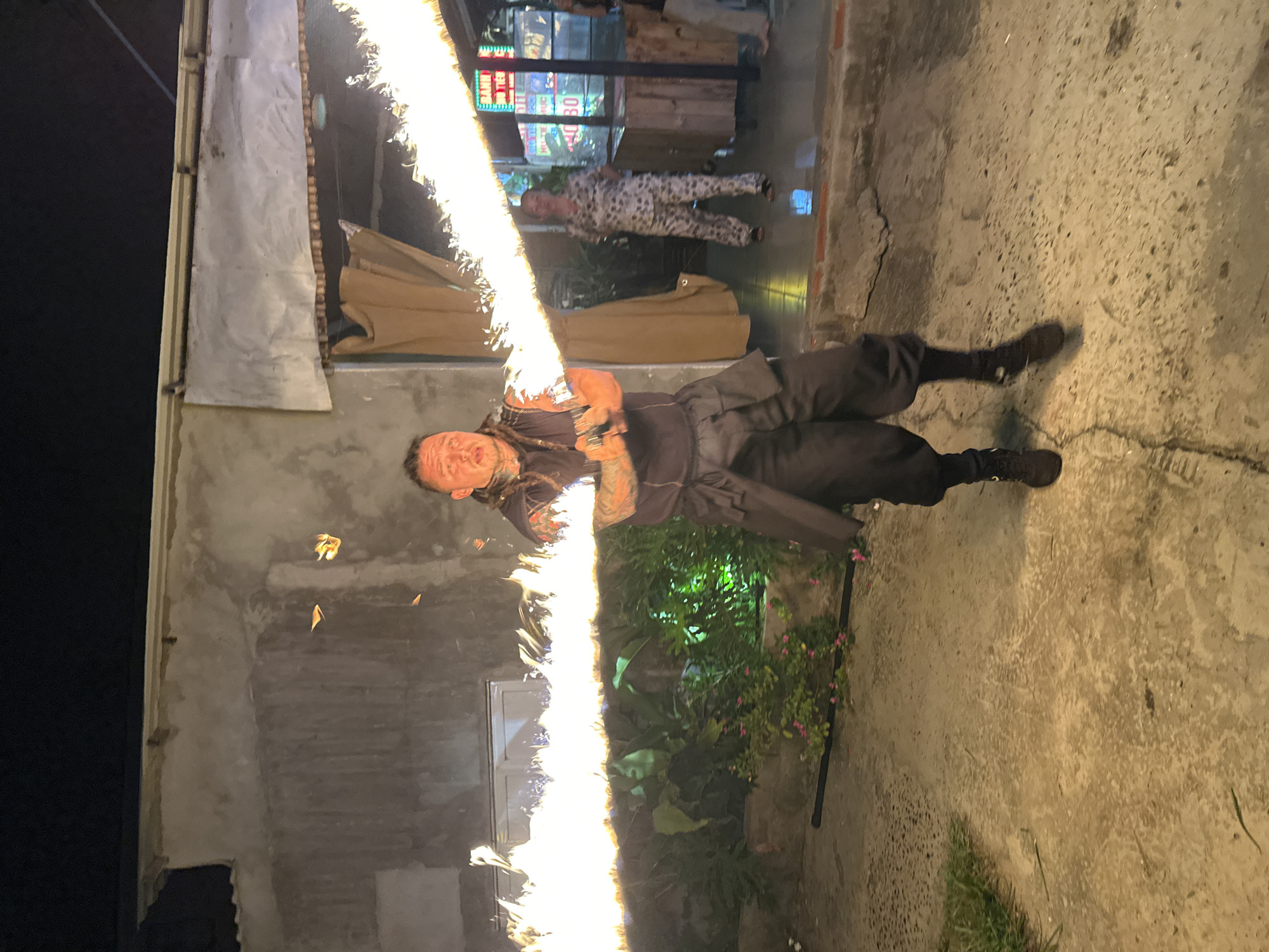 A male performer with dreadlocks and tattoos on his arms is performing fire breathing in an outdoor area at night. He is wearing a dark sleeveless shirt, dark baggy pants, and dark shoes. He is holding a fire-breathing apparatus that is emitting a long stream of fire horizontally across the image. In the background, there is a building with a stucco exterior, plants, and a woman in pajamas standing near a wooden counter. There are also some colorful signs visible in the background. The ground is made of concrete. The overall scene appears to be in Phu Thuy Ward, Vietnam, likely a tourist area given the street performance and signage.