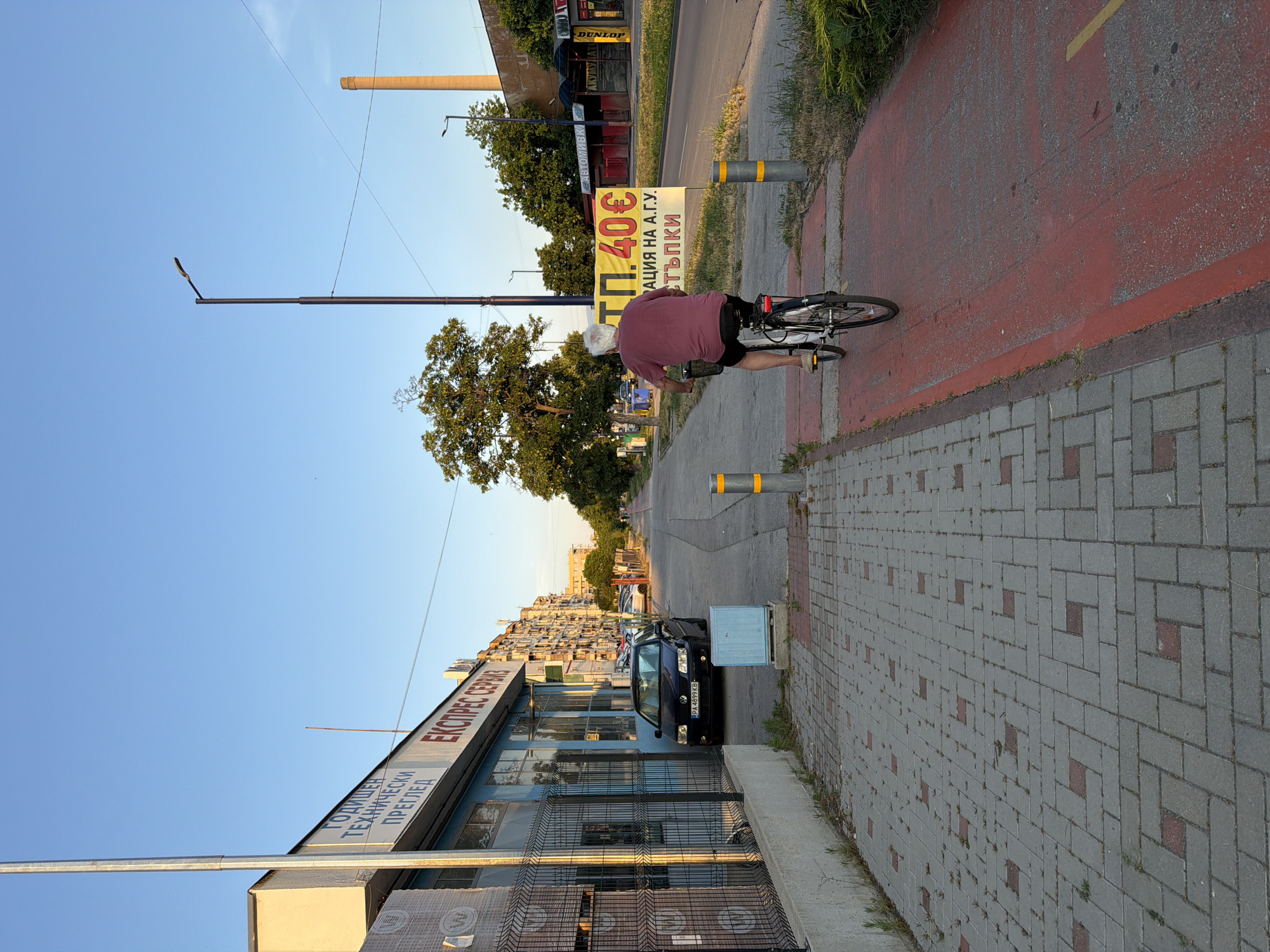The image depicts a man riding a bicycle on a red-marked bike lane next to a sidewalk in Pazardzhik, Bulgaria. The man is wearing a maroon t-shirt and black shorts, with gray hair. He is riding away from the camera towards the right side of the image. The sidewalk to the left of the bike lane is paved with grey and red interlocking bricks. Adjacent to the sidewalk is a building with blue walls and large windows, featuring signage with Bulgarian text. A black Volkswagen car is parked in front of the building. The environment includes a street with traffic in the background, trees, and buildings lining the street. A yellow sign with Bulgarian text and a price of "40€" is visible on the right side of the road. The sky is clear and blue, indicating a daytime scene. The overall impression is an urban setting on a sunny day.