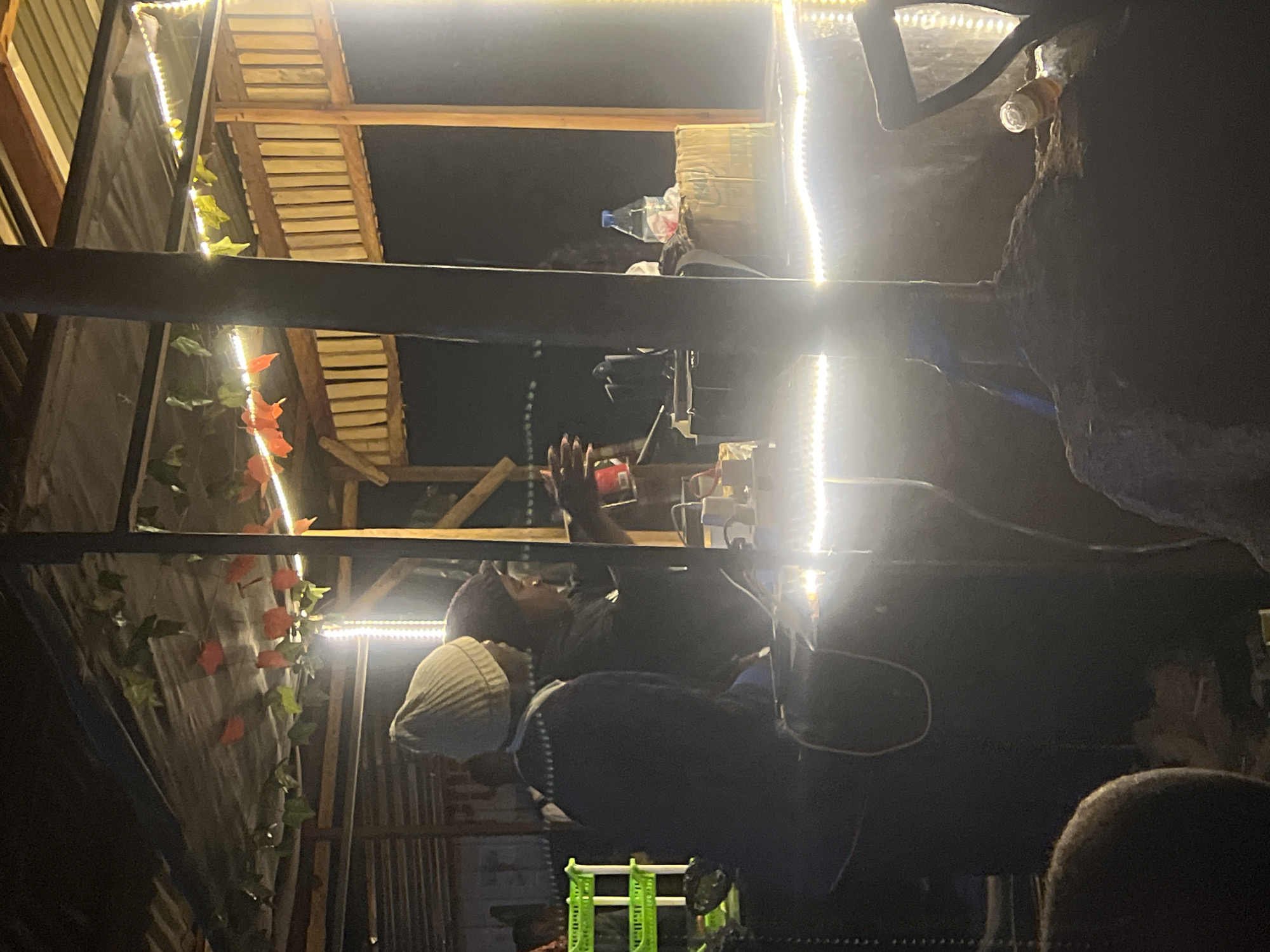 The image depicts an outdoor food stall at night in Durumi, Nigeria. Two individuals are visible, likely vendors, attending to customers or preparing food. The stall is illuminated by strings of white LED lights that highlight the wooden structure and roof, which is covered with a dark tarp decorated with artificial ivy and red flowers. In the background, a green plastic shelf unit is visible, and various items, including a bottle of water and cardboard boxes, are present on the counter. The overall scene suggests a lively, albeit dimly lit, nighttime market environment.
