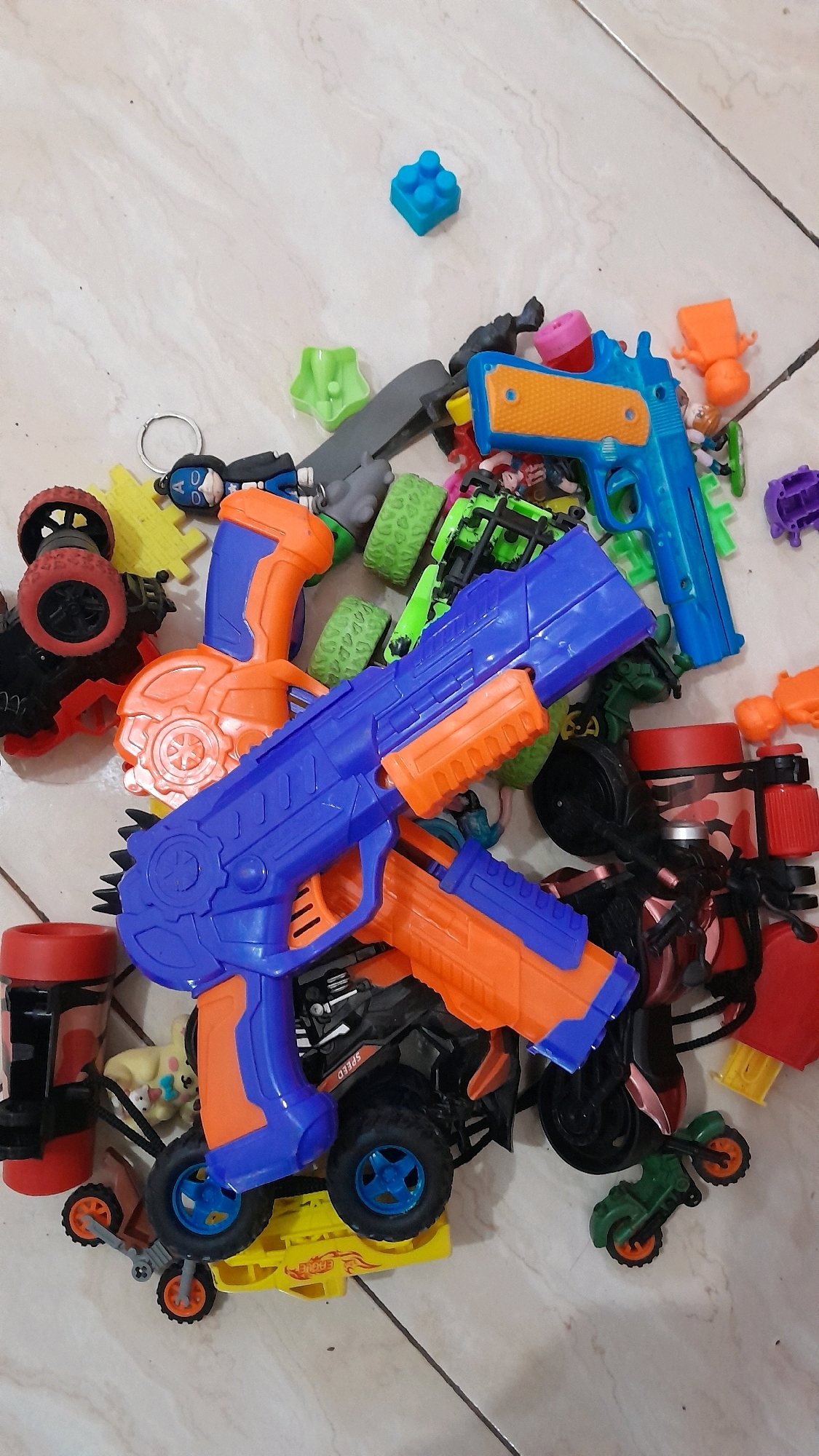 A top-down, eye-level shot captures a collection of colorful plastic toys scattered across a tiled floor. The arrangement is haphazard, with various toy vehicles and a toy gun prominently featured. A blue and orange toy gun is positioned in the foreground, angled across the frame. Behind it, another, smaller orange and blue toy gun lies on top of a green and black toy truck. Surrounding these are other toy vehicles, including a red toy car with large wheels, a yellow toy truck or car, and a small green toy motorcycle. Among the toys are also several other plastic objects in various shapes and colors, including a bright blue building block and a green star-shaped object. The floor is tiled with light-colored, square tiles, some of which show faint brown lines. The lighting is even, suggesting an indoor setting. The overall impression is a jumbled assortment of children's toys. The image does not contain any information that would allow for a determination of its location in Kentong, Indonesia.