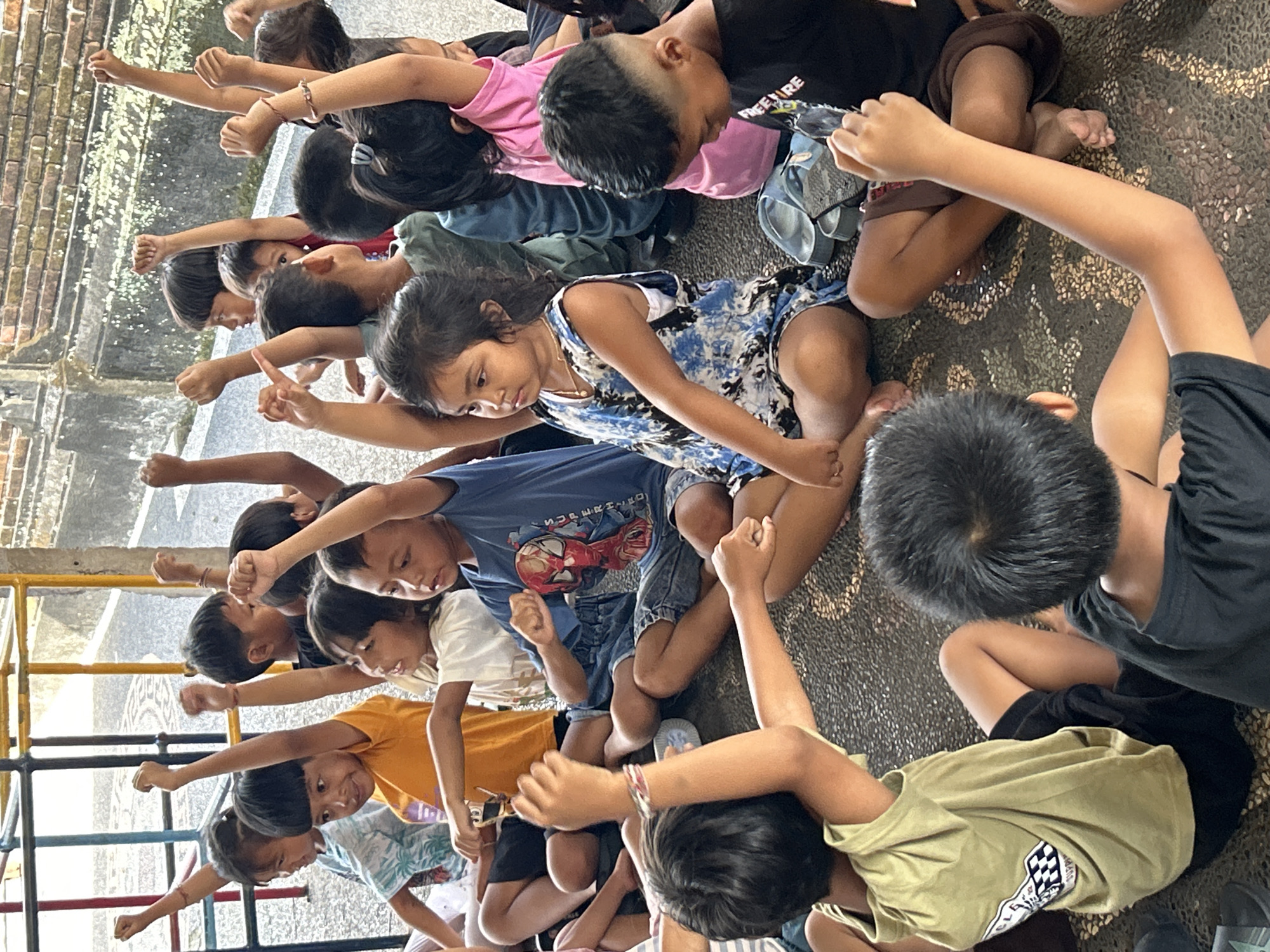 A group of children sits on a textured surface in Kediri, Indonesia. They are arranged in a semi-circular formation, all facing a similar direction. Each child has one arm extended upward, with their fist clenched. The children are wearing various casual clothes, including t-shirts and shorts. A metal structure is visible in the background, suggesting an outdoor setting. The light indicates that it is likely daytime. The children's focused expressions and gestures suggest a coordinated activity or game.