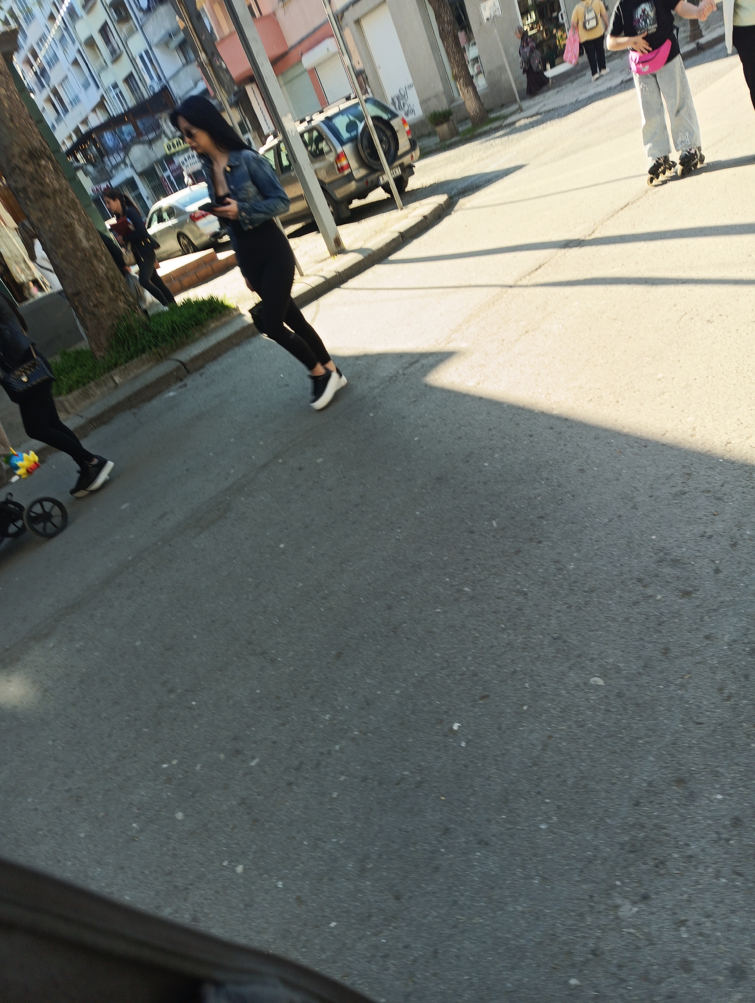 The photo depicts an urban scene likely in Burgas, Bulgaria, judging by architectural styles. The photograph is taken at street level, capturing part of a road and adjacent sidewalks. Several individuals are visible; one is walking and looking at her phone. A person on roller skates is visible on the right side of the image. A car and a few pedestrians appear in the background. The environment includes buildings, trees, and the paved road surface. Shadow patterns suggest strong sunlight. A stroller is on the sidewalk to the left.