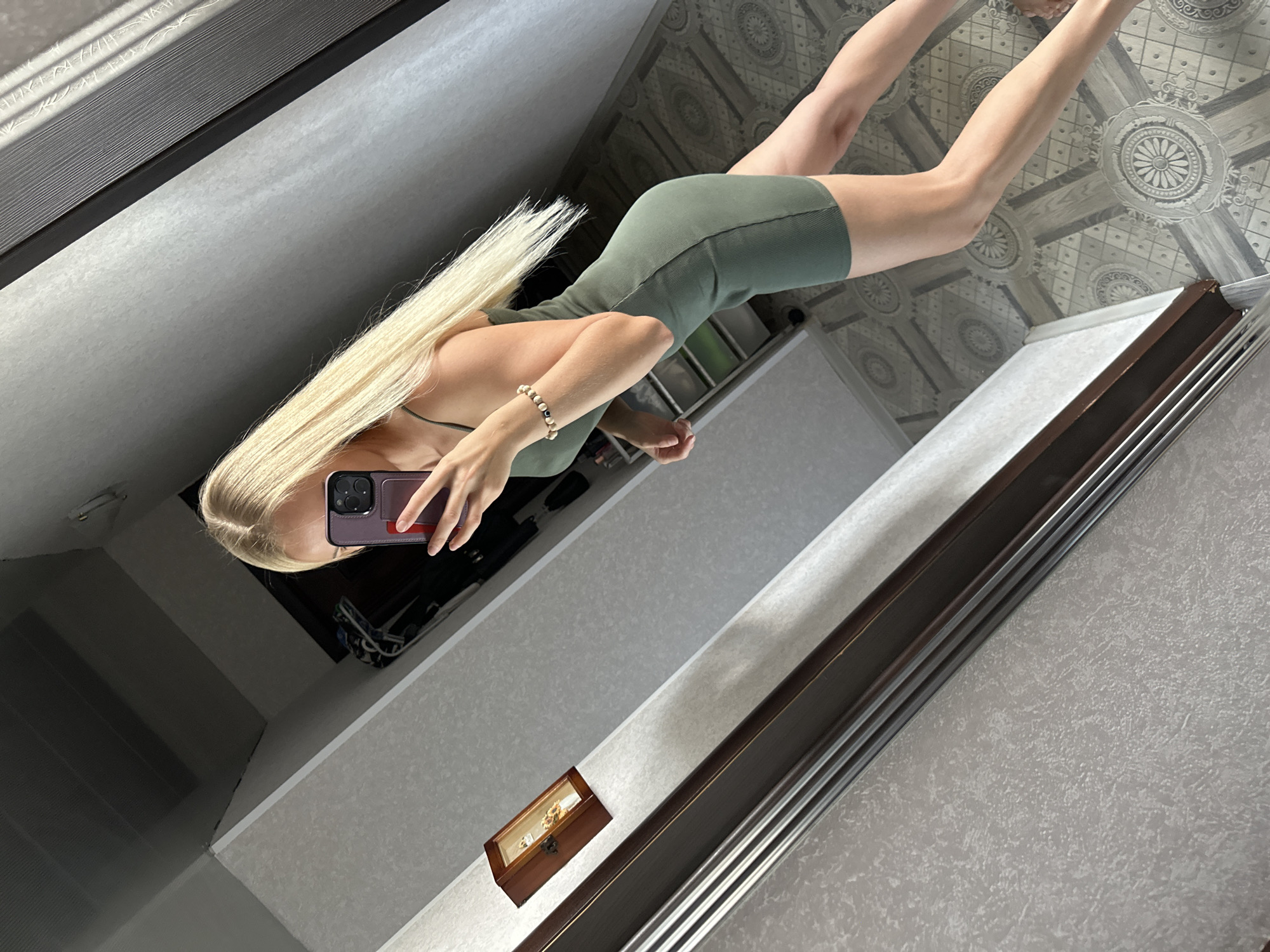 A blonde woman is taking a mirror selfie in a room. She is wearing a form-fitting olive green dress and a bracelet on her left wrist. Her hair is long and straight. The room appears to be a bedroom or dressing area, with patterned wallpaper and a dark wooden closet or dresser visible in the reflection. The floor has an ornate, circular pattern. A small wooden framed object containing small figures is hanging on the wall to the left. The image is taken from a slightly elevated, angled perspective. The scene is consistent with an interior residential space in Energetik, Russia.