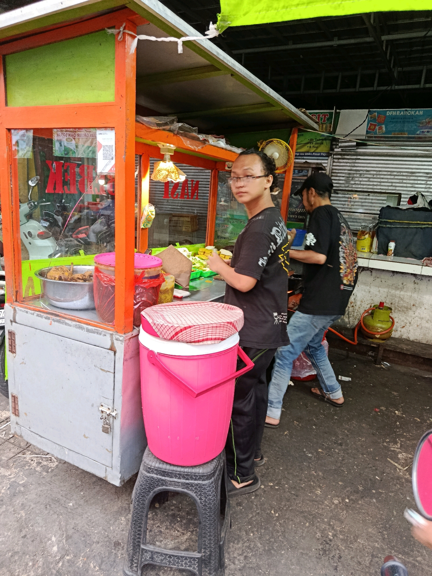 A man wearing glasses and a black t-shirt stands in front of an orange and green food stall in Bekasi, Indonesia. He has a neutral expression. Behind him, another person wearing a black t-shirt and blue jeans is preparing food. The food stall is stocked with various items, including large containers of food and drinks. A pink cooler is placed on a black stool to the left of the stall. The ground is asphalt, and there are some puddles and debris visible. The overall scene depicts a busy street food vendor operating in an urban environment.