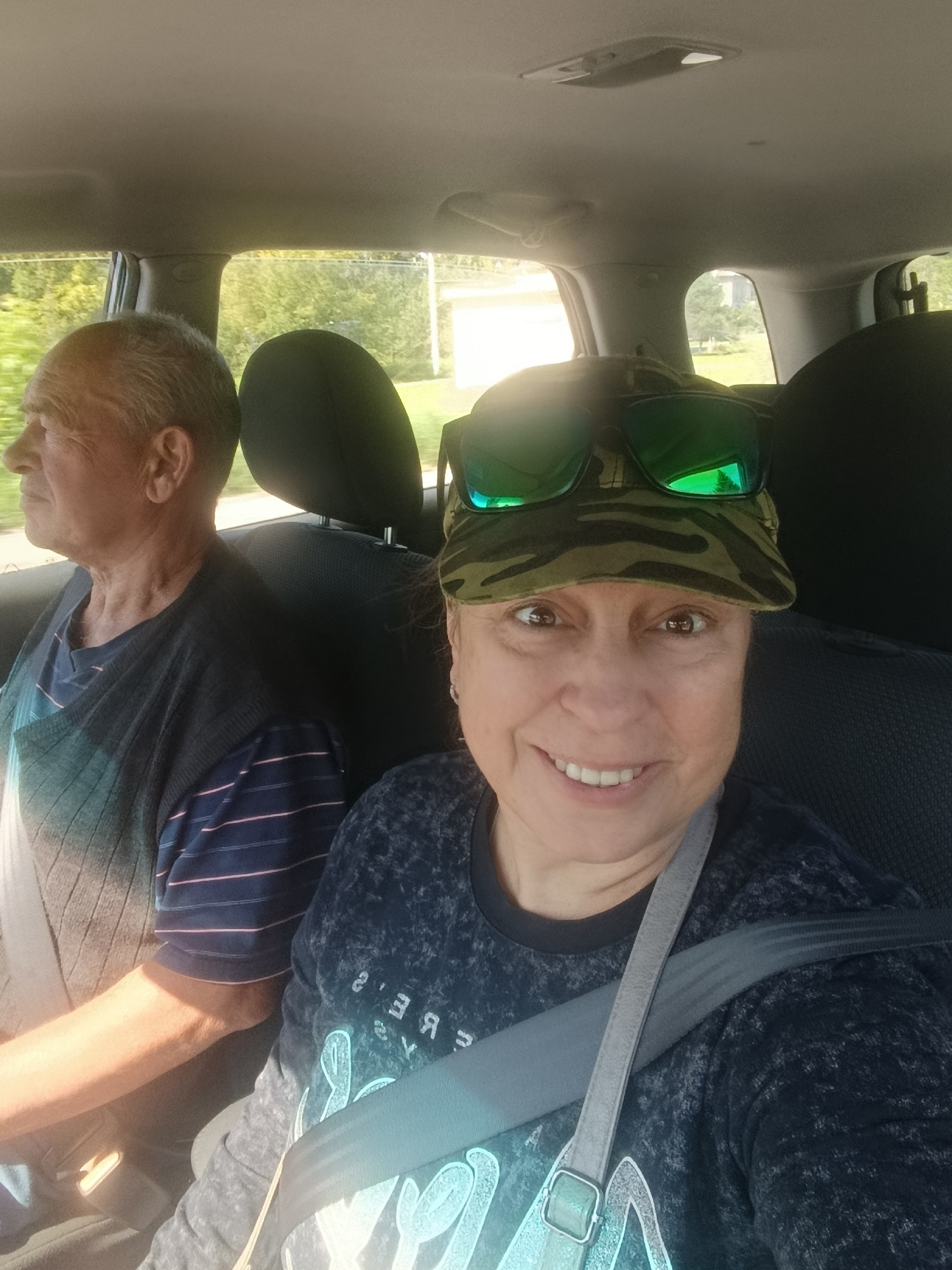 Two people are in the front seats of a vehicle, appearing to be traveling. A man is seated on the left, looking out the side window, wearing a dark sweater over a striped t-shirt. A woman is seated on the right, facing the camera and smiling. She is wearing a camouflage baseball cap with large green-lensed sunglasses perched on top. Her t-shirt has a distressed texture and a graphic print on the front. Both individuals are wearing seatbelts. The background visible through the car windows suggests a rural or suburban setting with greenery and trees. The overall lighting indicates it is daytime. The image is a selfie taken from the perspective of the woman. There is no specific information about the location in the city of Снопок Новый, Russia, visible in the image.