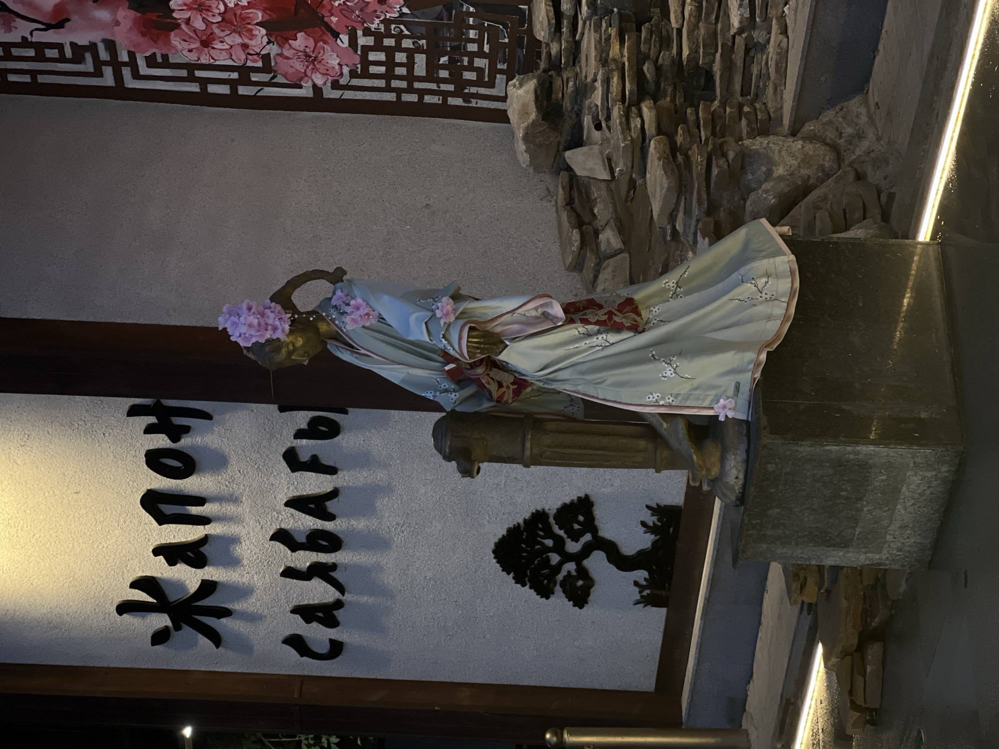 The image shows a statue of a woman in a traditional Japanese kimono, adorned with pink flowers, standing in front of a building. The building features a sign with Cyrillic script that reads "Жапон Саябағы," which translates to "Japanese Garden" in English. To the left of the statue, a silhouette of a bonsai tree is visible on the wall. To the right, a decorative wooden lattice and a depiction of pink cherry blossoms are present. The area around the statue is landscaped with stones and illuminated by warm lighting, suggesting an outdoor space. This scene is located in the city of Shymkent, Kazakhstan, likely at an establishment named or themed after a Japanese garden.