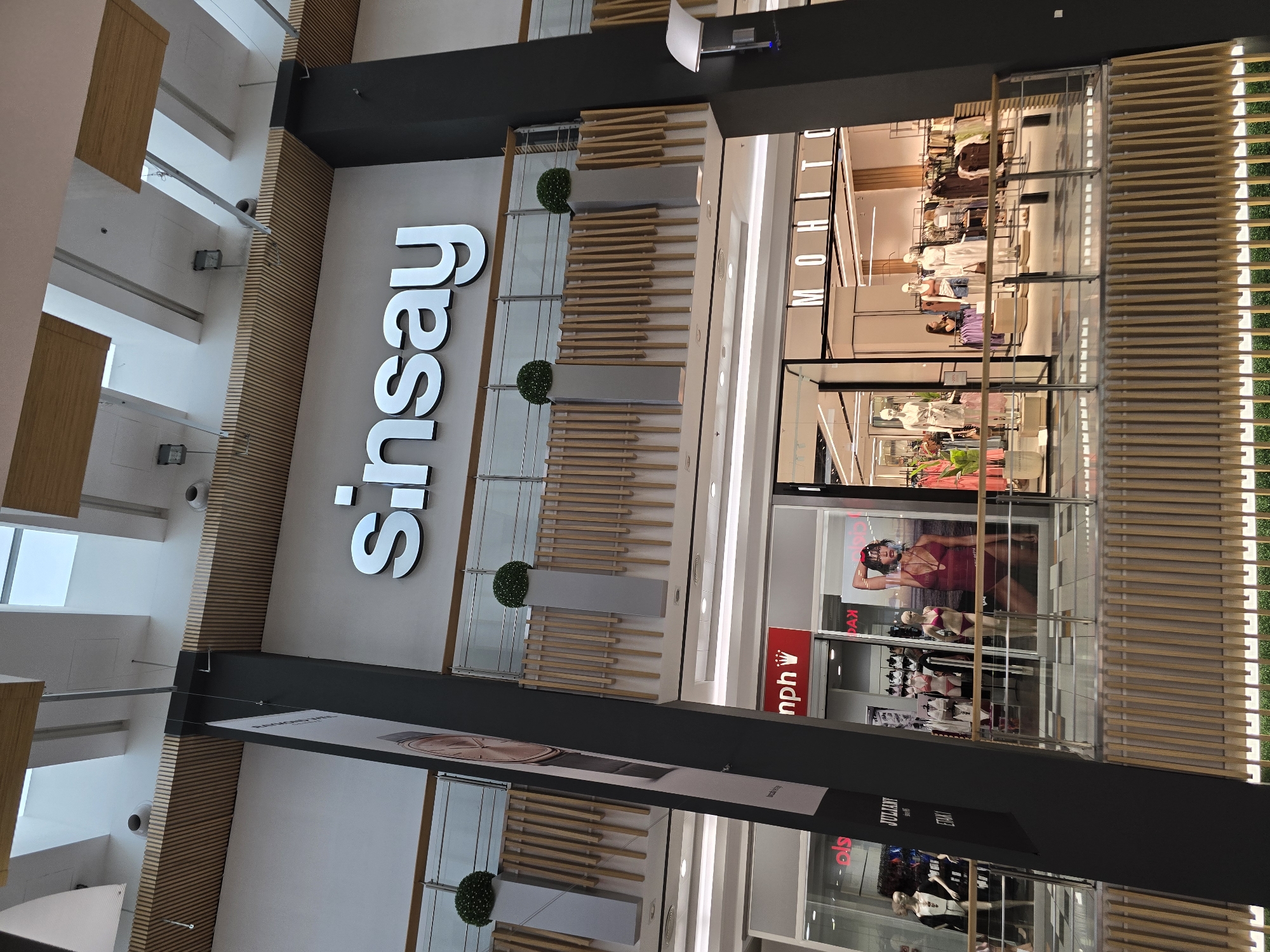 The image shows the interior of a shopping mall in Plovdiv, Bulgaria. The prominent signage "Sinsay" is visible at the top of the image, indicating a clothing store. Below, other store facades are visible, including "Mohito," and a store with "nph" and a crown logo, likely "Triumph." Several mannequins are displayed in the store windows, dressed in various outfits, including swimwear. The mall features a modern design with a high ceiling, wooden paneling, and balconies. The lighting is bright, typical of an indoor retail environment. No people are clearly identifiable in the image, though some figures appear to be present inside the stores, possibly staff or shoppers.