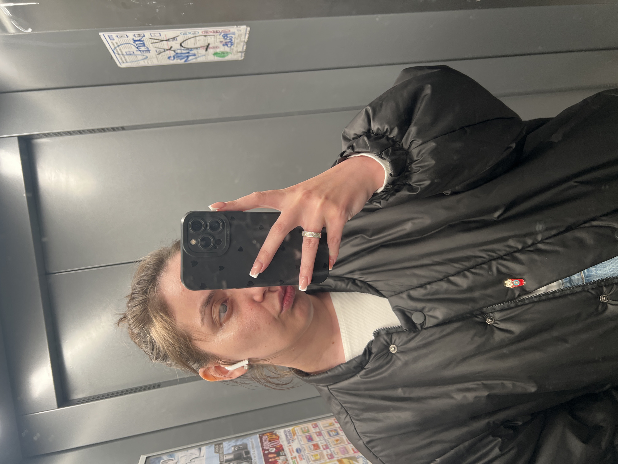 A woman is taking a selfie in what appears to be an elevator. She is wearing a black puffer jacket and has an earbud in her ear. She is holding her phone up to her face. There is a sticker on the wall behind her and another image is partly visible below her. The environment is metallic, appearing to be the interior of an elevator. The location cannot be determined.