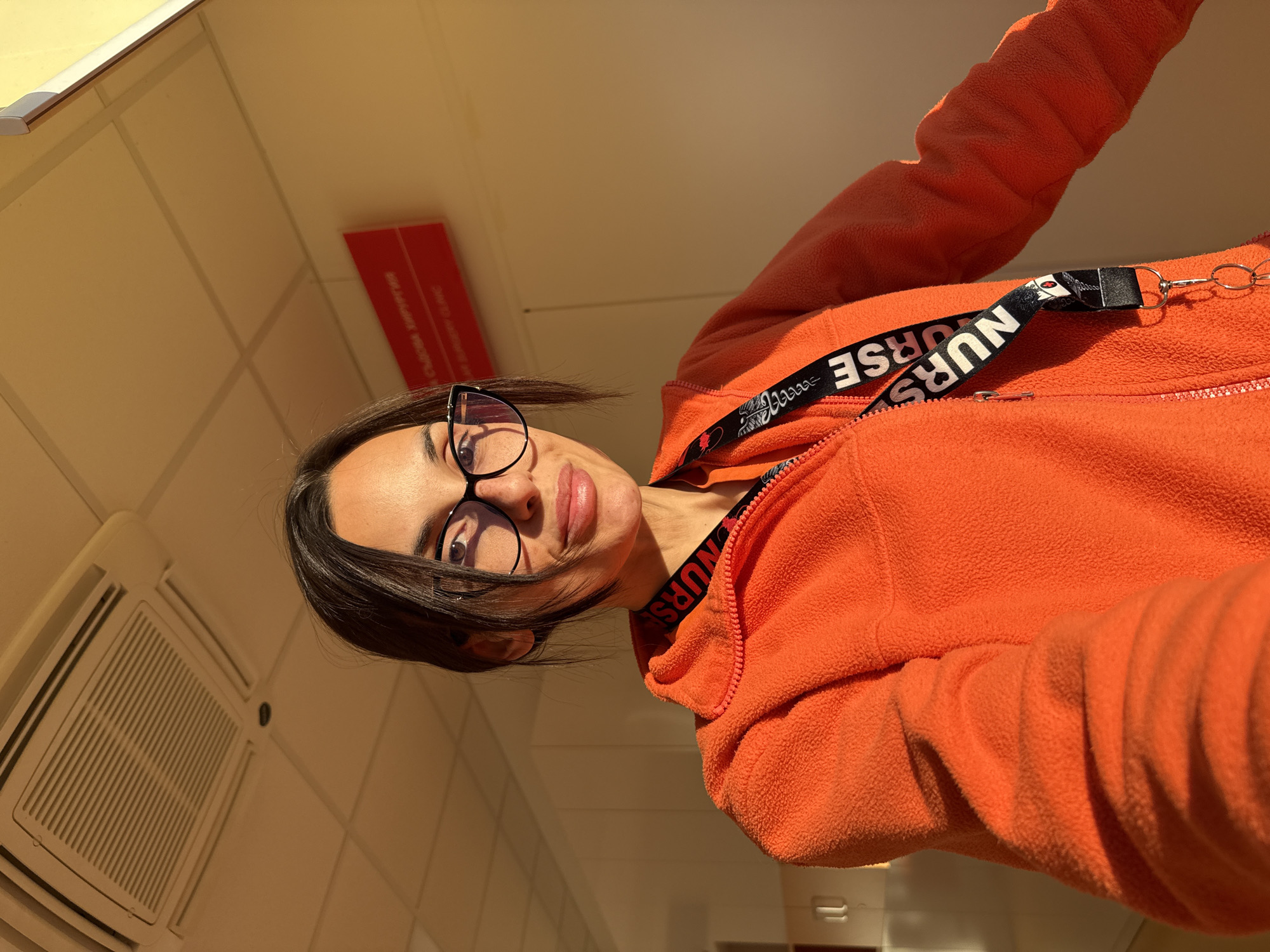 A woman with brown hair and glasses is wearing an orange fleece jacket with a black lanyard that says "NURSE." The photo is taken from a low angle, looking up at the woman. The background shows a ceiling with a ventilation system, tiled walls, and a red sign with text in Bulgarian. The sign likely indicates a location in Pleven, Bulgaria, given the city was specified.