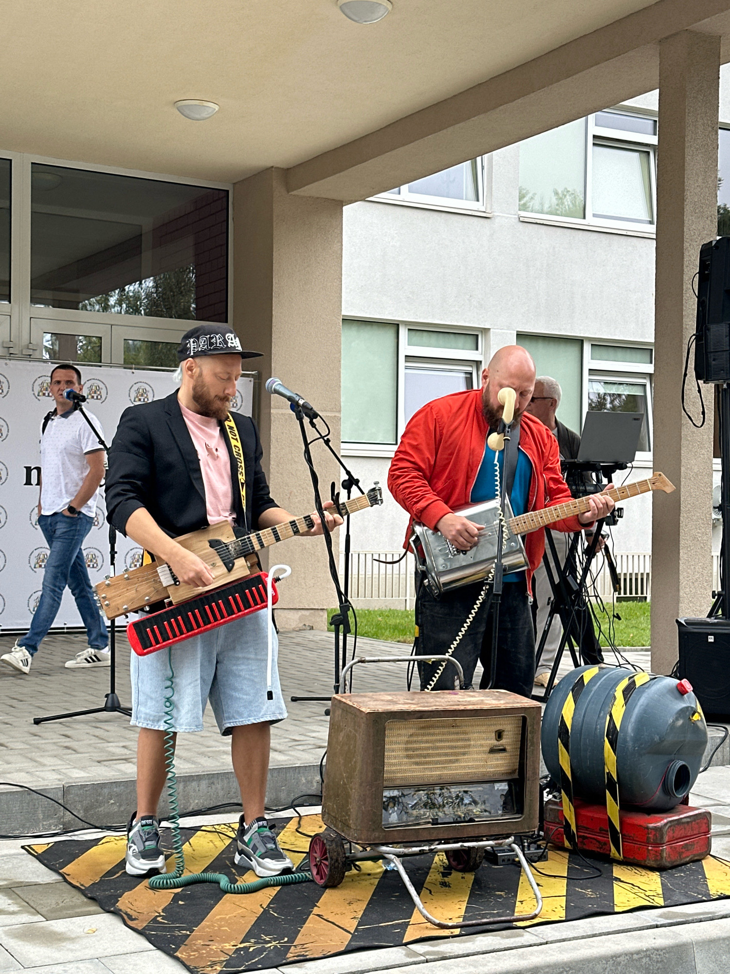The photograph captures an outdoor musical performance on a paved area in front of a modern building with large windows. Two men are actively playing musical instruments that appear to be custom-made or modified. The man in the foreground, wearing a black jacket, pink t-shirt, and light blue shorts, is playing a wooden instrument that resembles a guitar with an attached red keyboard. He is also wearing a black baseball cap. To his right, another man in a red jacket and blue shirt is playing a metal instrument that looks like a toaster with a guitar neck attached, and he has a phone receiver near his mouth as a microphone. Behind them, another man stands near a microphone, and further back, a person is visible near a laptop on a stand. In the foreground, a vintage radio sits on a cart, next to a large grey container with yellow and black hazard stripes. The building in the background suggests an institutional or office setting, possibly a school or public facility. The scene suggests a performance event taking place outdoors.