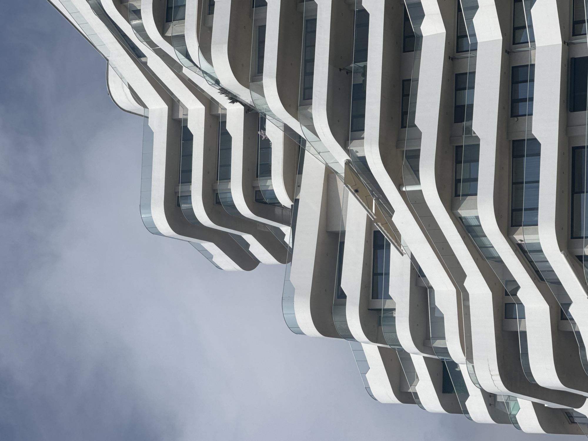 The image shows an abstract upward view of a modern building in Burgas, Bulgaria. The building features a white facade with repeating vertical curved sections that create a wave-like pattern. Each curved section contains glass balconies with clear railings. Windows are visible behind the curved sections. The sky is visible in the background and to the left of the building. There is a person standing on one of the balconies.