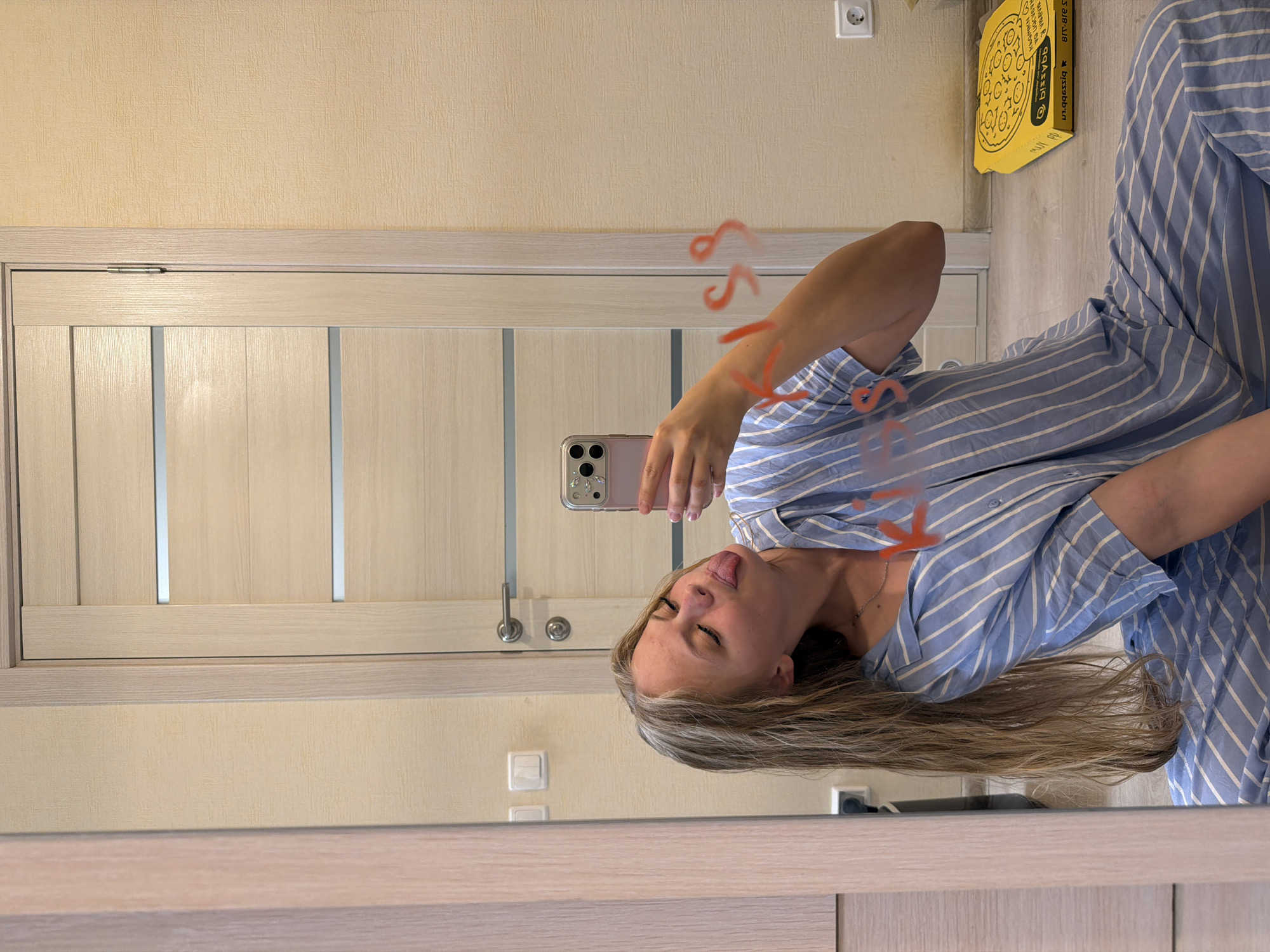 The image shows a young woman taking a mirror selfie in what appears to be a residential setting, possibly a bedroom or hallway in Izhevsk, Russia. The woman is wearing a blue and white striped pajama top. She is holding a phone, which is reflecting in the mirror. Her eyes are closed and she is sticking out her tongue, conveying a playful or mischievous emotion. Red scribbles, possibly drawn on the mirror, are visible on her arm and torso.
In the background, a light-colored door with vertical paneling is visible, along with a textured yellow wall and a light-colored skirting board. On the wall, there's a white electrical outlet and a bright yellow pizza box with a circular design and some text. The lighting suggests it is daytime.