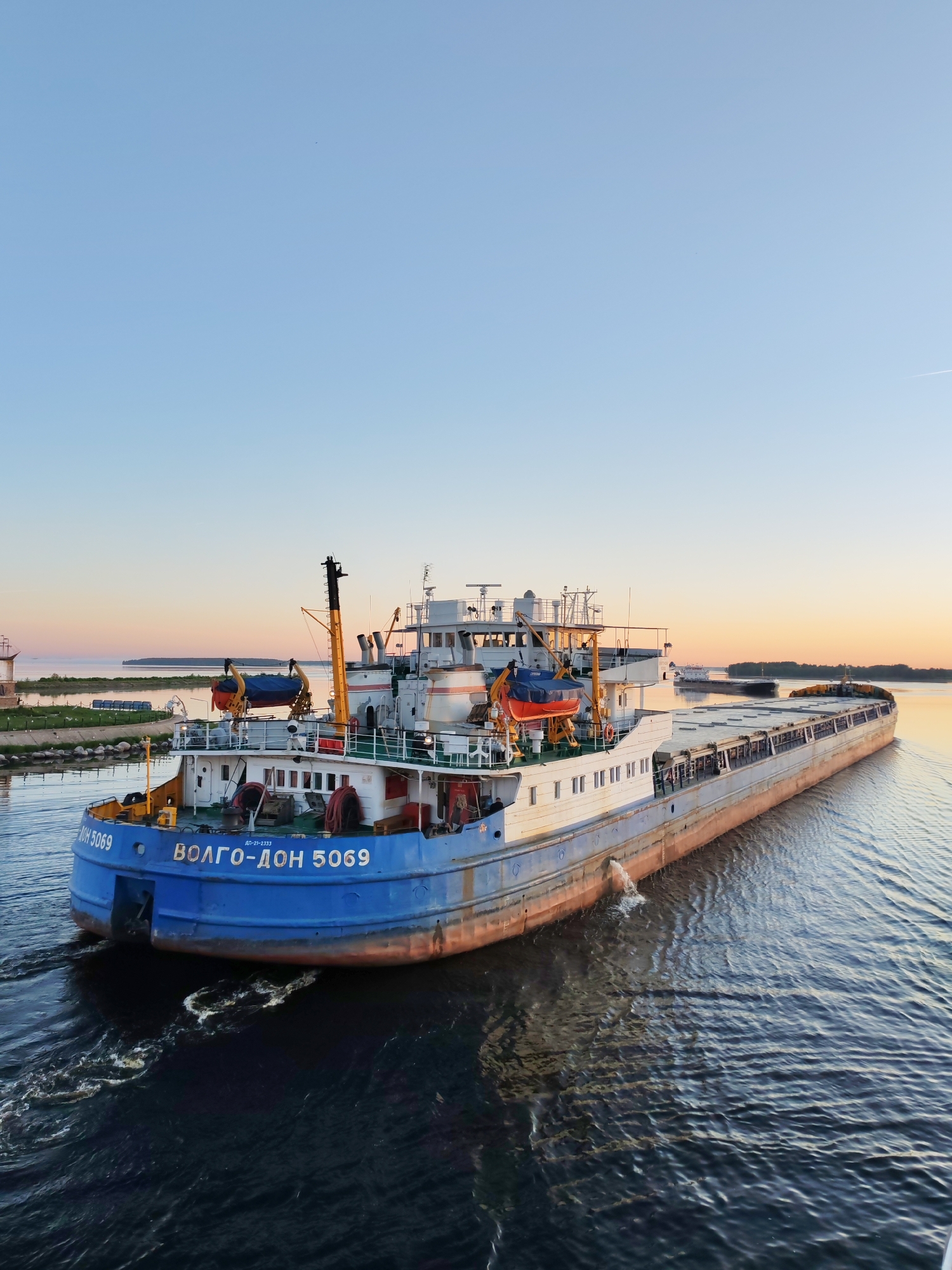 A long, blue and white cargo vessel, identified by the Cyrillic inscription "ВОЛГО-ДОН 5069" (VOLGO-DON 5069) on its stern, navigates a wide body of dark blue water during what appears to be either sunrise or sunset. The sky transitions from a light, clear blue overhead to a pale orange-yellow at the horizon. The ship's superstructure, situated at the stern, is white and features multiple windows, railings, and what appears to be a radar mast and various antennae. Two yellow davits are visible; one holds an orange life raft covered with a blue tarp. The main body of the ship is a long cargo area with gray hatch covers, and the lower part of its hull is painted a reddish-brown. A distinct wake of churning white water trails behind the moving vessel, and a stream of water is actively being discharged from a pipe on its starboard side. On the left side of the frame, a green grassy bank with some low-lying structures is visible, with a distant landmass or island beyond it. Another faint landmass with trees is present on the right horizon. No individuals are clearly visible on the ship or in the surrounding environment.