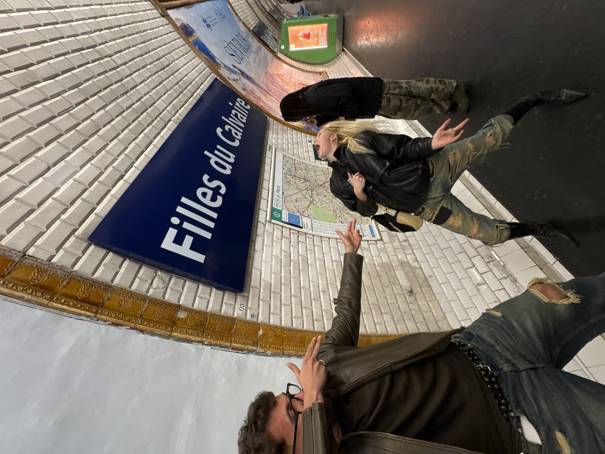 The image shows a group of people in the Paris metro station "Filles du Calvaire." A person with curly dark hair and glasses is in the foreground, wearing a brown leather jacket and blue jeans with rips. This person's right hand is raised. Behind them, a blonde woman in a black leather jacket and camouflage pants is lying on the ground with her mouth open. Another person wearing a black jacket and camouflage pants is further in the background. A metro map and advertisements are visible on the wall. The walls are tiled in white, with a decorative brown border. A green vending machine is visible in the background.