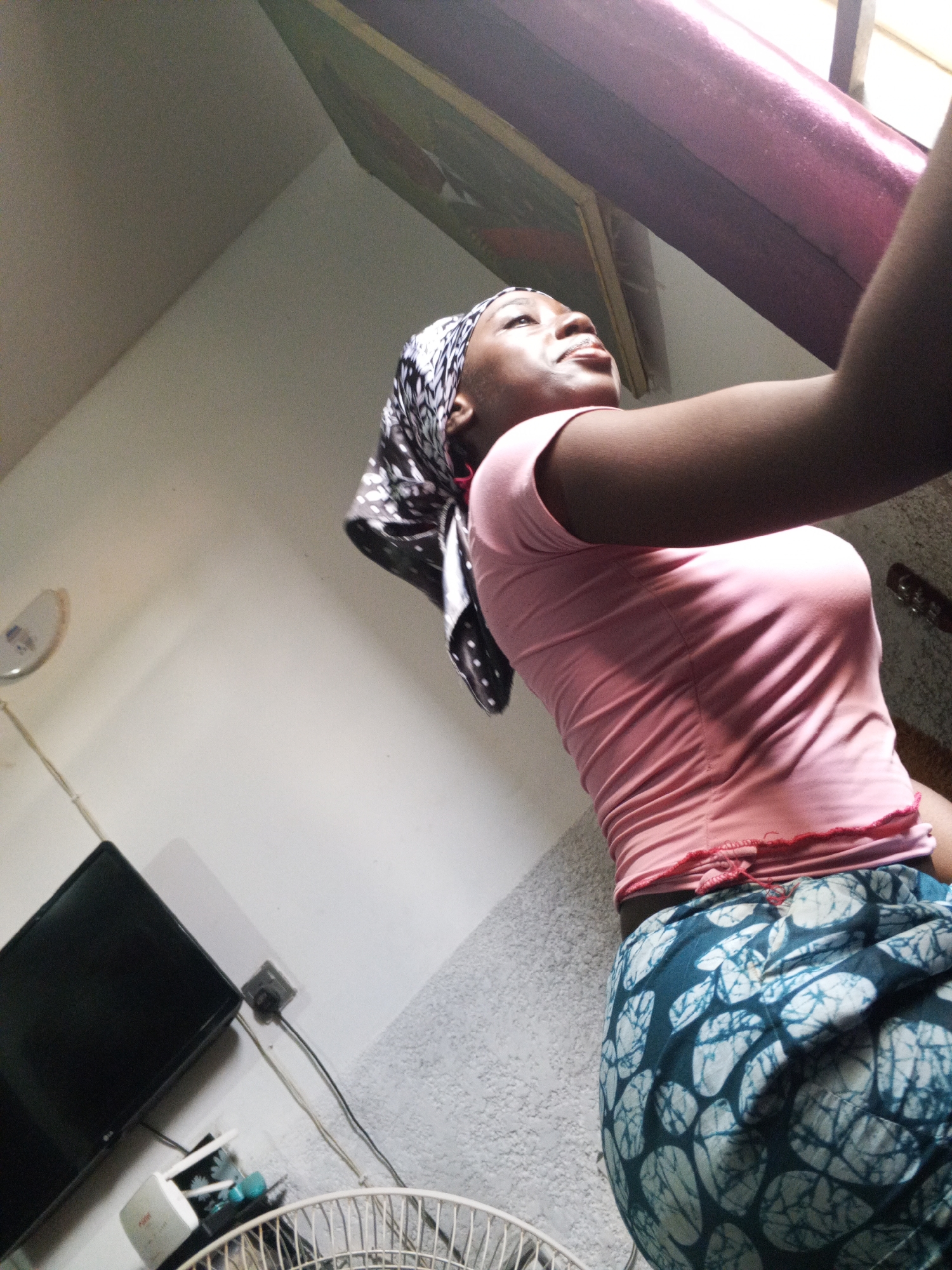 A young woman stands indoors, possibly in a private residence. She wears a pink T-shirt, a patterned blue and white skirt, and a headscarf with a black and white pattern. Her gaze is directed upward, possibly looking out a window. Visible in the background are a television set, a wall-mounted electrical outlet, a floor fan, and what appears to be a framed picture or artwork. The lighting in the room seems to be a combination of natural light from the window and artificial light. The location is possibly in a residential area of Abuja, Nigeria, considering the attire of the woman, the interior style, and the electrical outlet type.