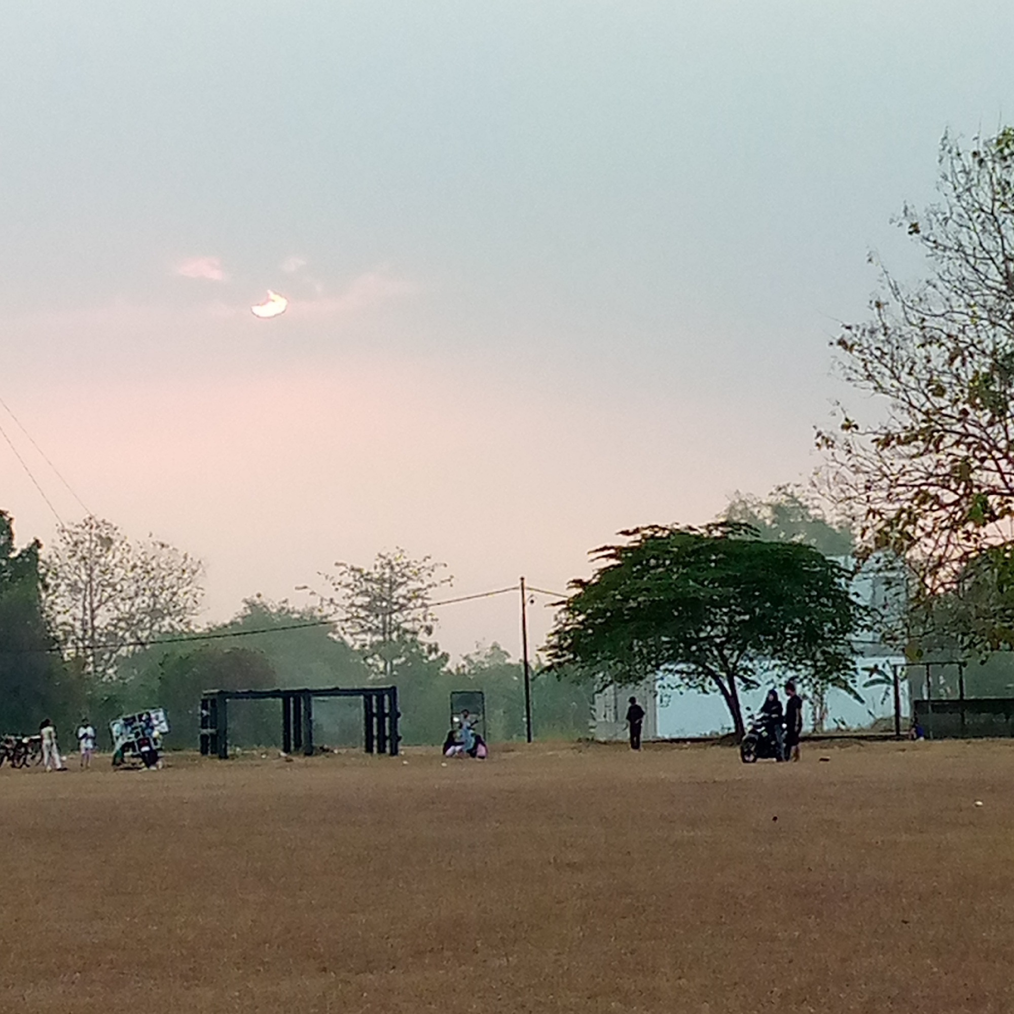 The image depicts an outdoor scene at what appears to be a public ground or field, possibly in Pacing, Indonesia. In the sky, a partial solar eclipse is visible, with the sun appearing as a crescent shape. The sky is a pale blue with a gradient of pinkish hues near the horizon, suggesting it might be early morning or late afternoon.
In the foreground, the ground is covered in dry, brown earth, possibly a sports field or a dusty area. Several individuals and objects are scattered across the scene. On the left, there are a few people standing near what appear to be motorcycles or bicycles. Further into the middle ground, there is a dark, rectangular structure, possibly a fence or a goalpost, with two more people standing near it. To the right of this structure, a large, leafy green tree stands prominently.
In the mid-to-far ground, there are more structures and a few more people. One group is gathered around a motorcycle or scooter. To the far right, a person is standing near a white building or wall. The overall atmosphere seems relatively calm, with a few people engaged in various activities. The presence of the partial solar eclipse is the most striking celestial event in the sky.